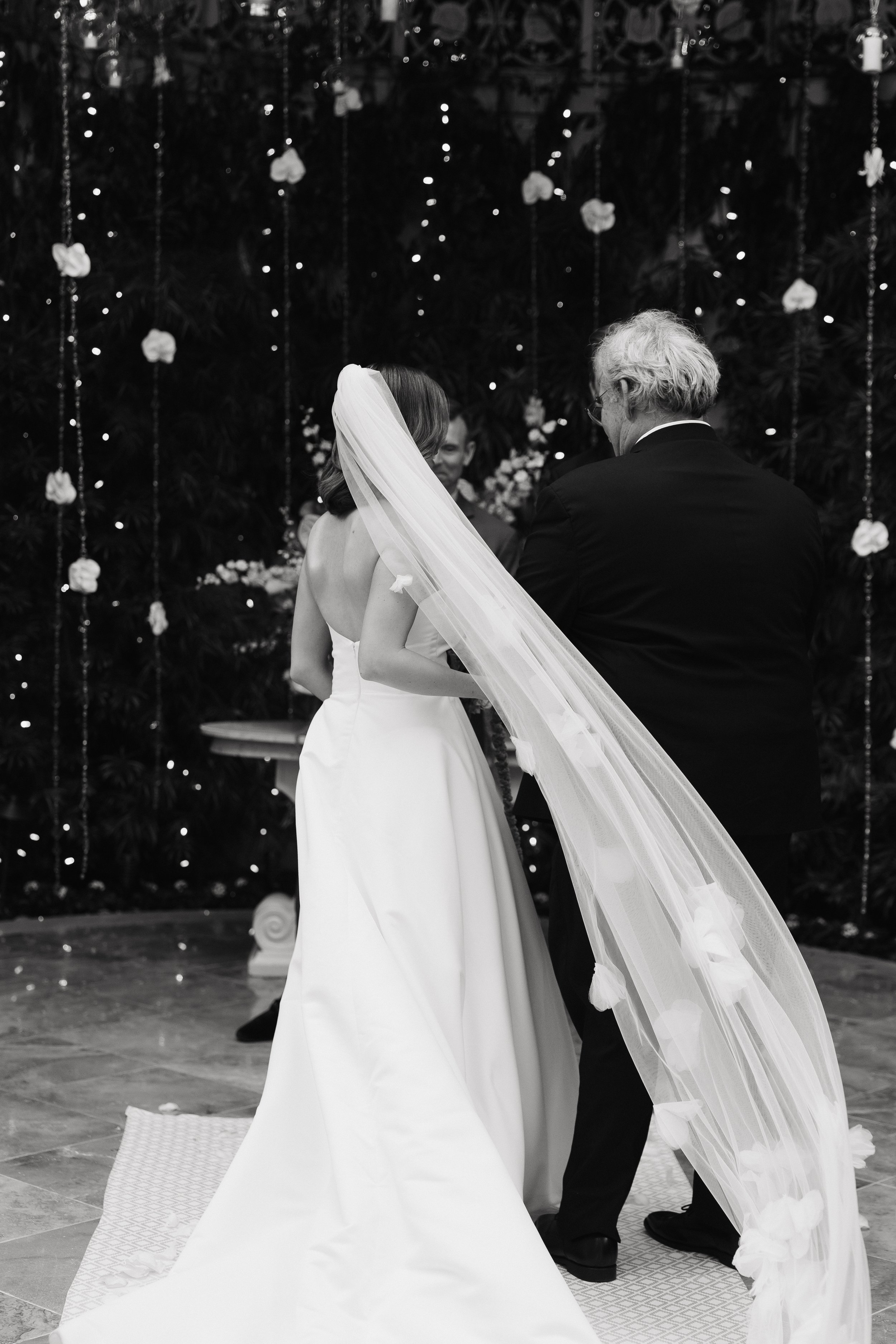 A bride and groom exchanging vows during a wedding ceremony, with the bride wearing a white wedding gown and veil, and the groom in a black suit, against a decorated backdrop of flowers and lights.