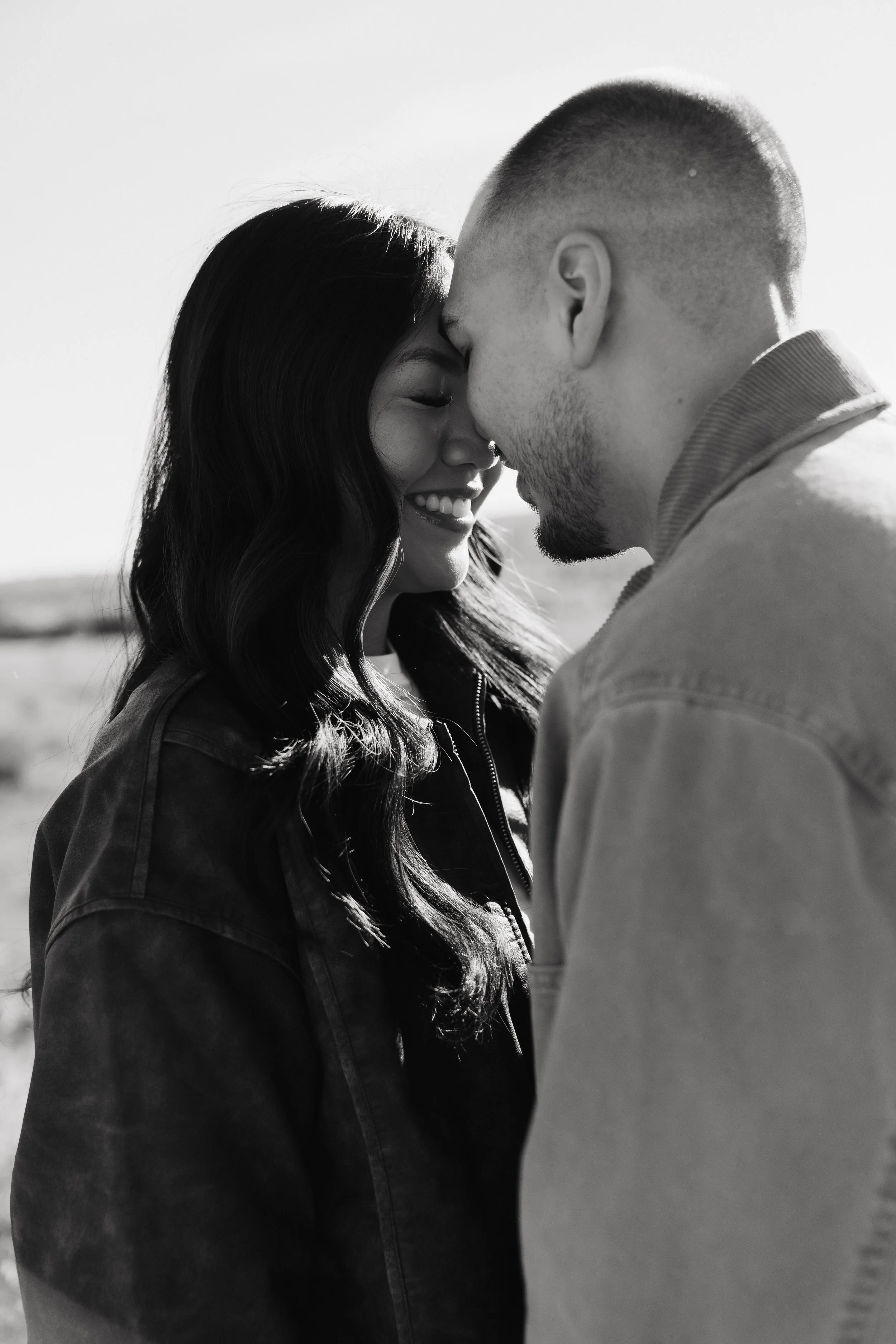 A black-and-white photo of a man and a woman with their foreheads touching, smiling and closing their eyes. The woman has long, wavy hair and is wearing a jacket. The man has short hair, facial hair, and is wearing a shirt with the collar visible.