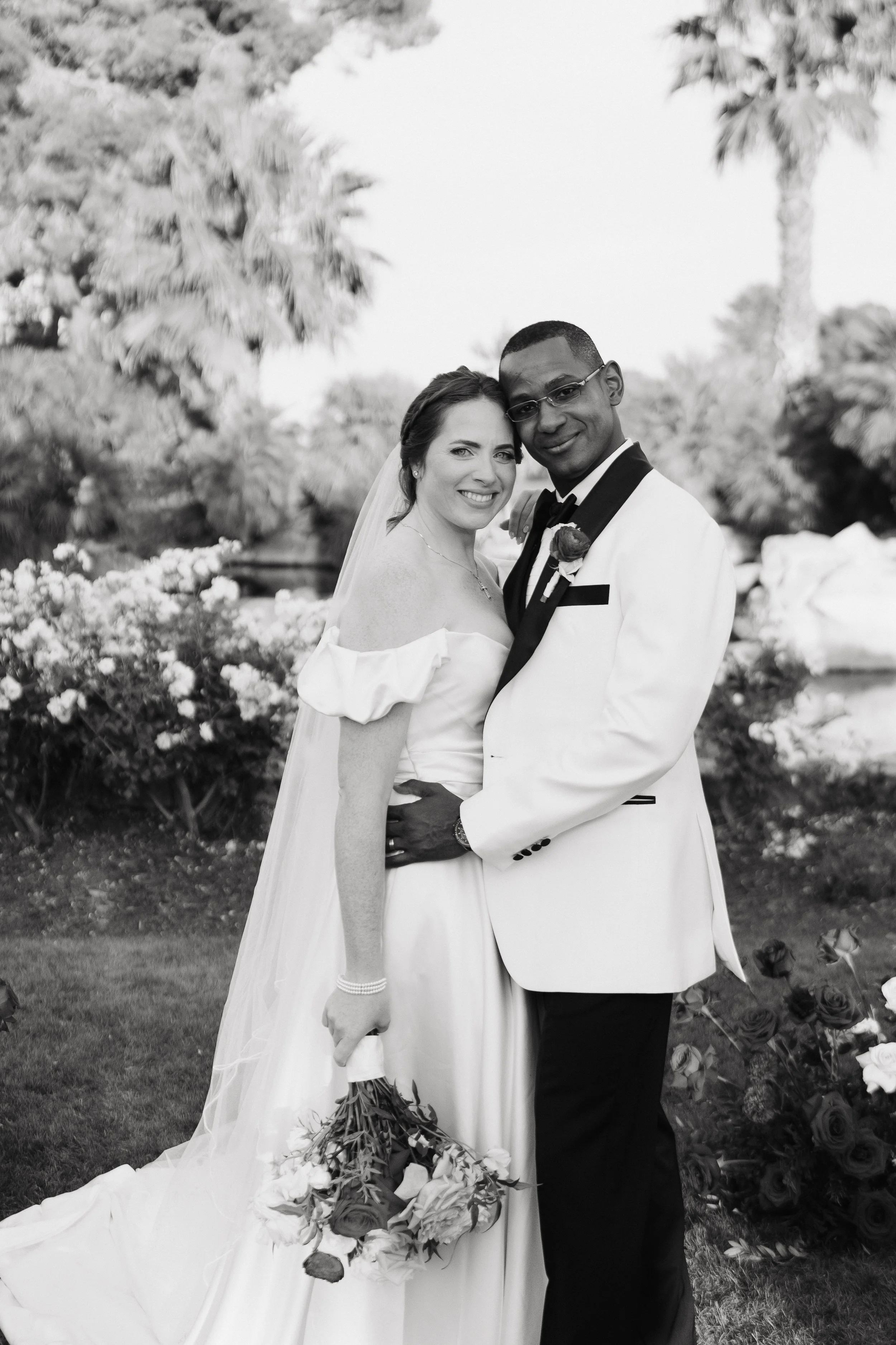 A black and white photo of a bride and groom smiling and posing outdoors, with palm trees and flowers in the background.