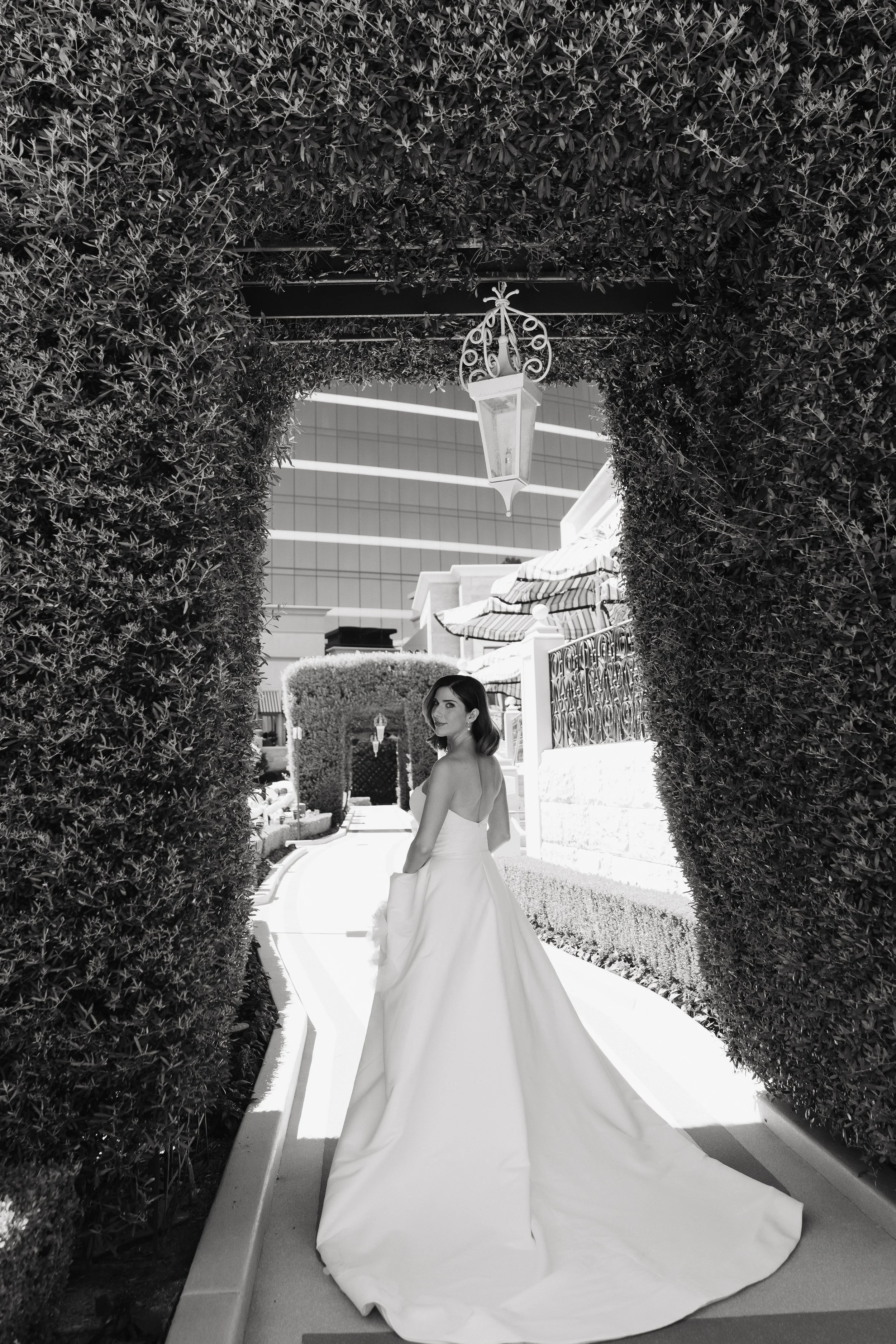 Black and white photo of a woman in a wedding dress standing beneath an arched opening decorated with dense hedges and a hanging lantern in an outdoor setting.