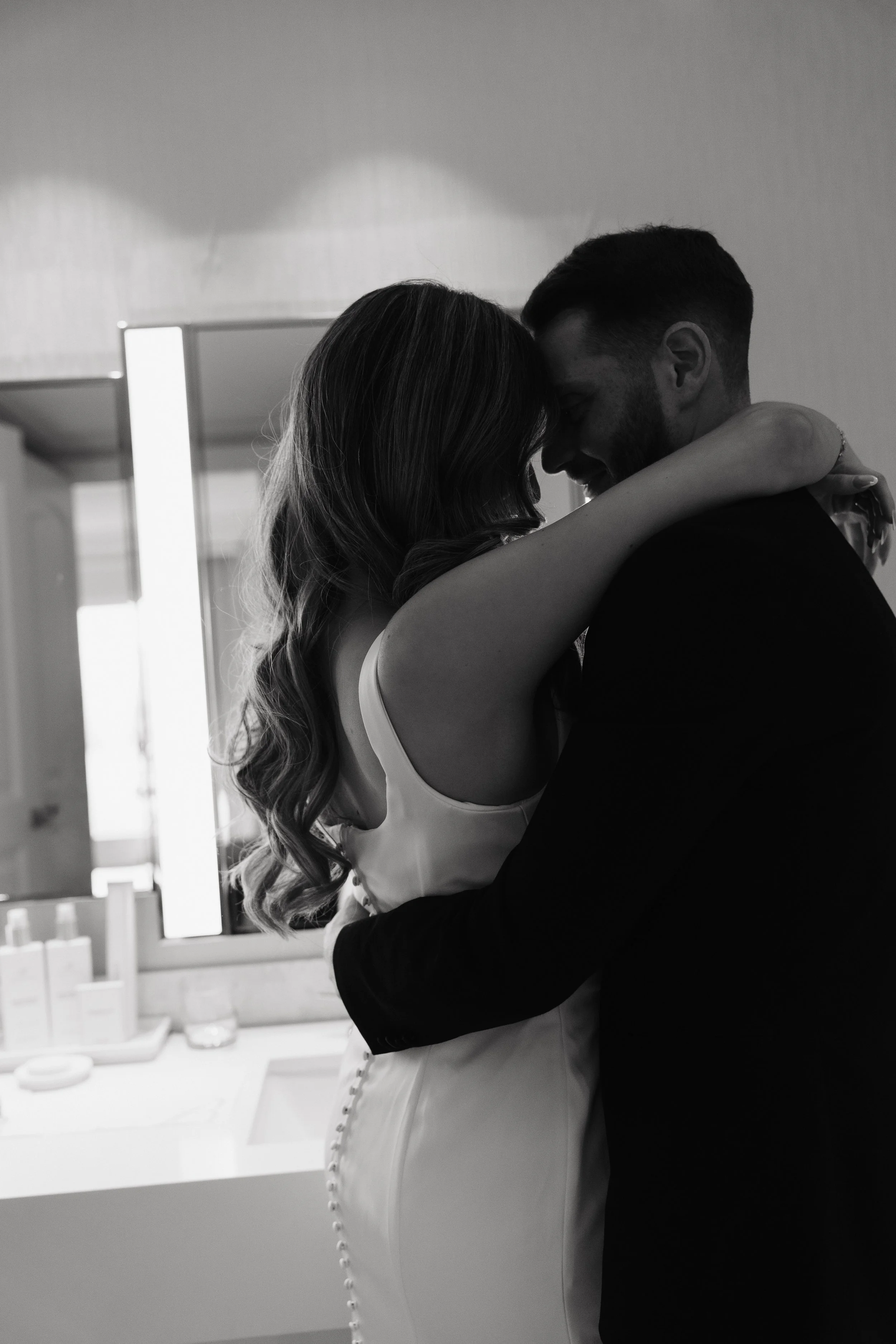 A black-and-white photo of a couple in an embrace, with their foreheads touching and eyes closed, in a room with a window and bathroom items in the background.