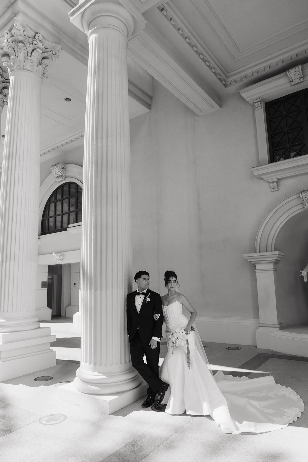 Wide black-and-white portrait of a bride and groom framed by grand columns and classical architecture.