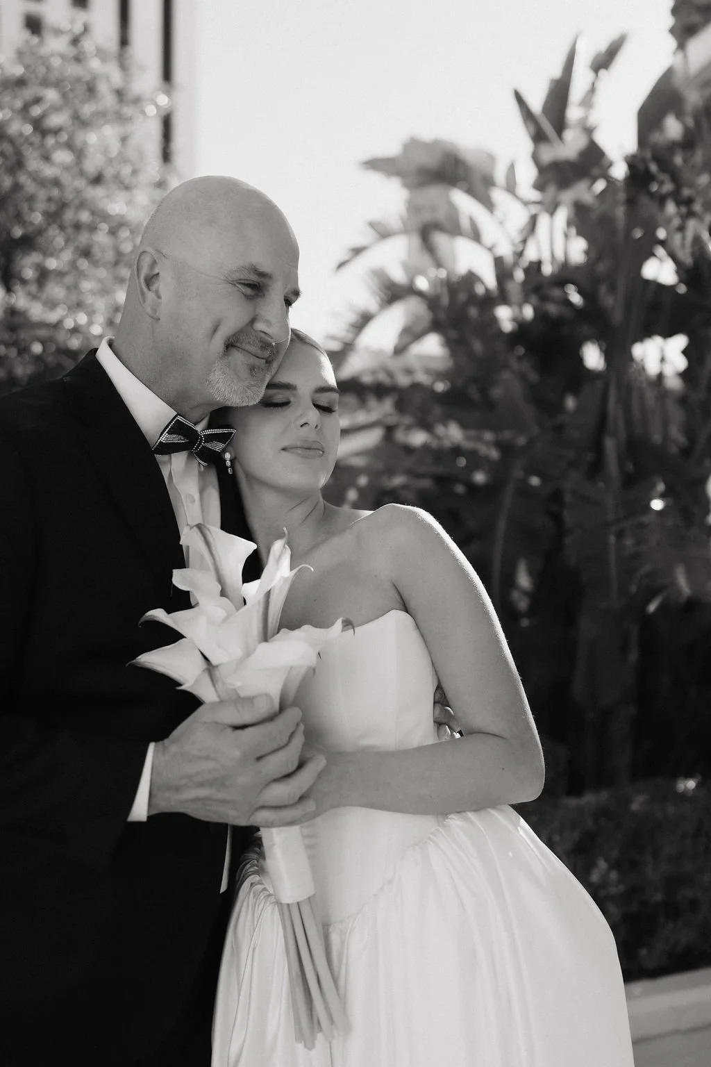 A black and white photo of a bride holding a bouquet of flowers with her eyes closed, leaning her head against a man, who is smiling with his eyes closed.