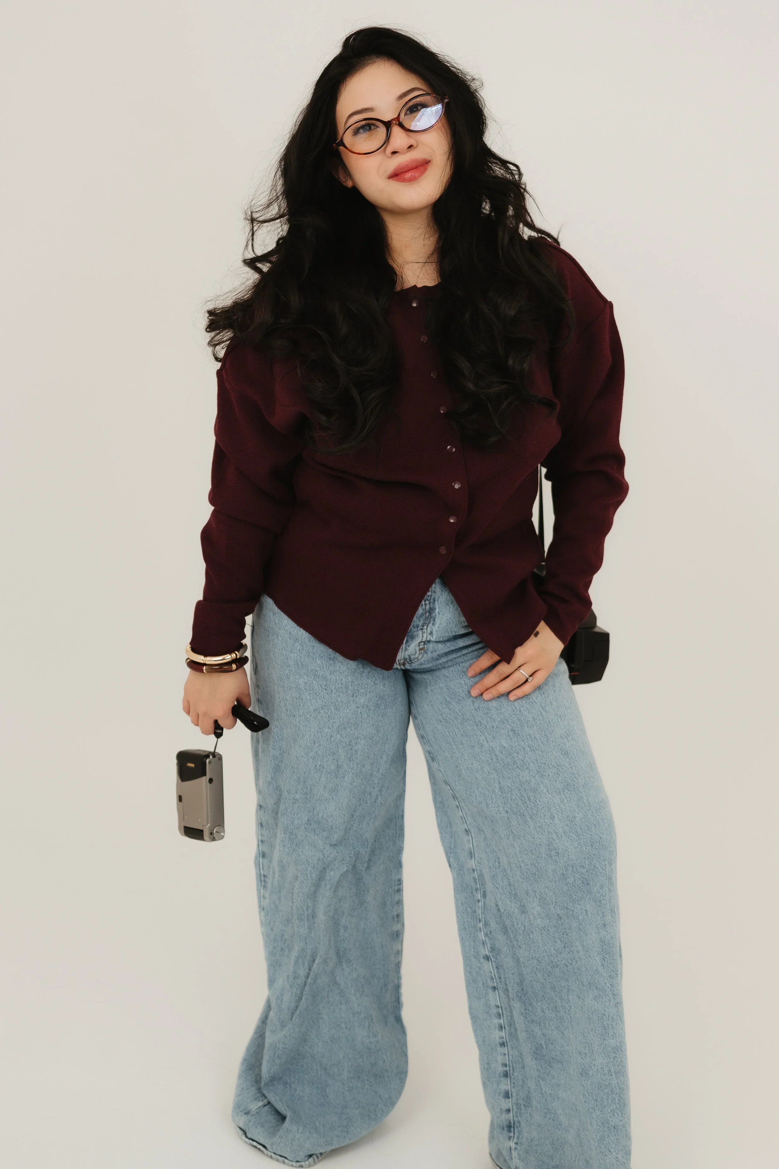 A Las Vegas photographer wearing a maroon shirt and light blue jeans, holding a camera in her right hand, standing against a plain white background.