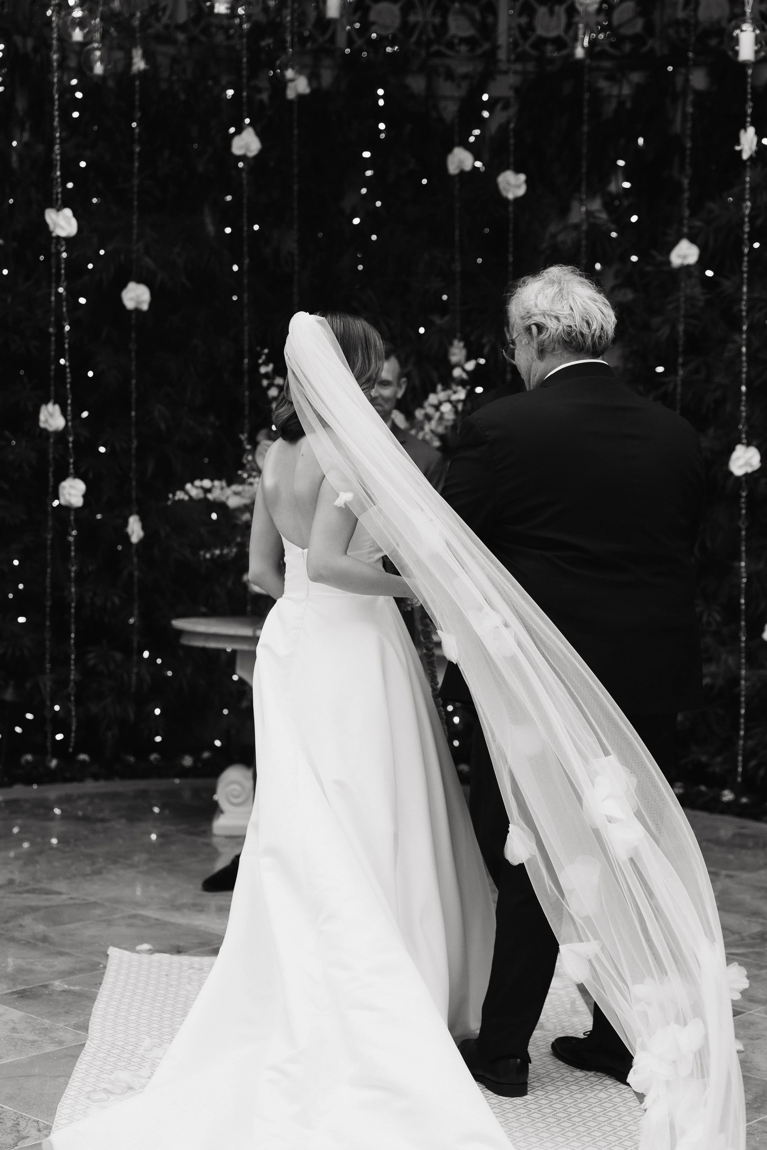 A black-and-white photo of a bride and her father during a wedding ceremony, with floral and fairy light decorations in the background.