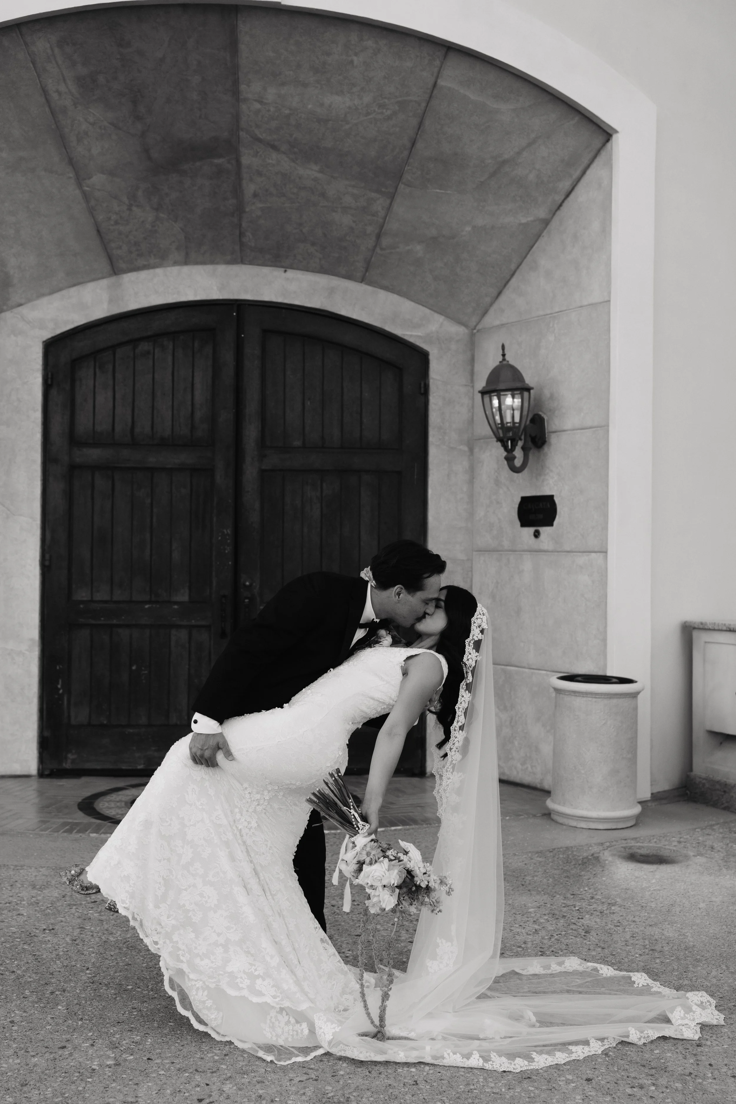 A bride and groom kissing outside a building, with the groom dipping the bride. The bride is holding a bouquet of flowers, and the scene includes a large wooden door, a lantern, and a stone column.