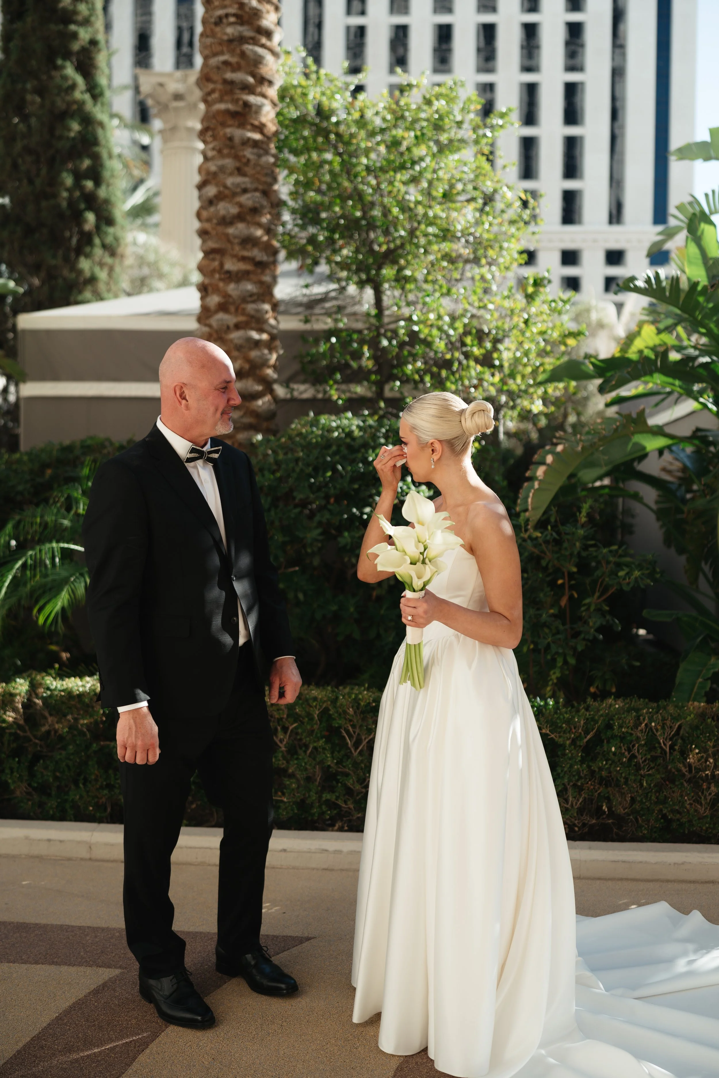 A bride with platinum blonde hair in a bun is holding white calla lilies and wiping away a tear, standing outside. A man in a black tuxedo with a bow tie is standing to her left, smiling gently. Green plants and trees, tall buildings, and a white col