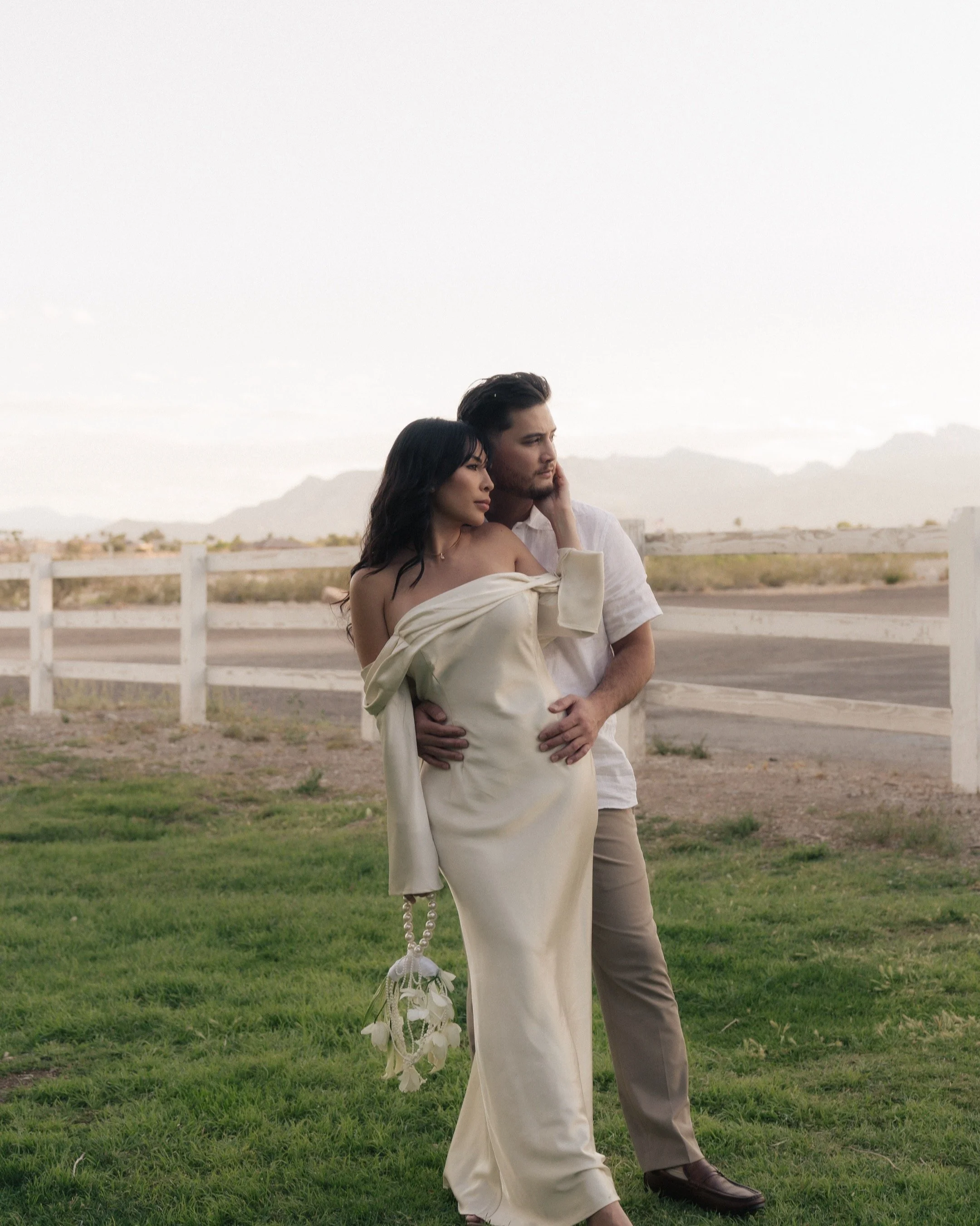 A man and woman stand close together outdoors in front of a white fence and distant mountains on a cloudy day, with the woman resting her head on the man's shoulder.