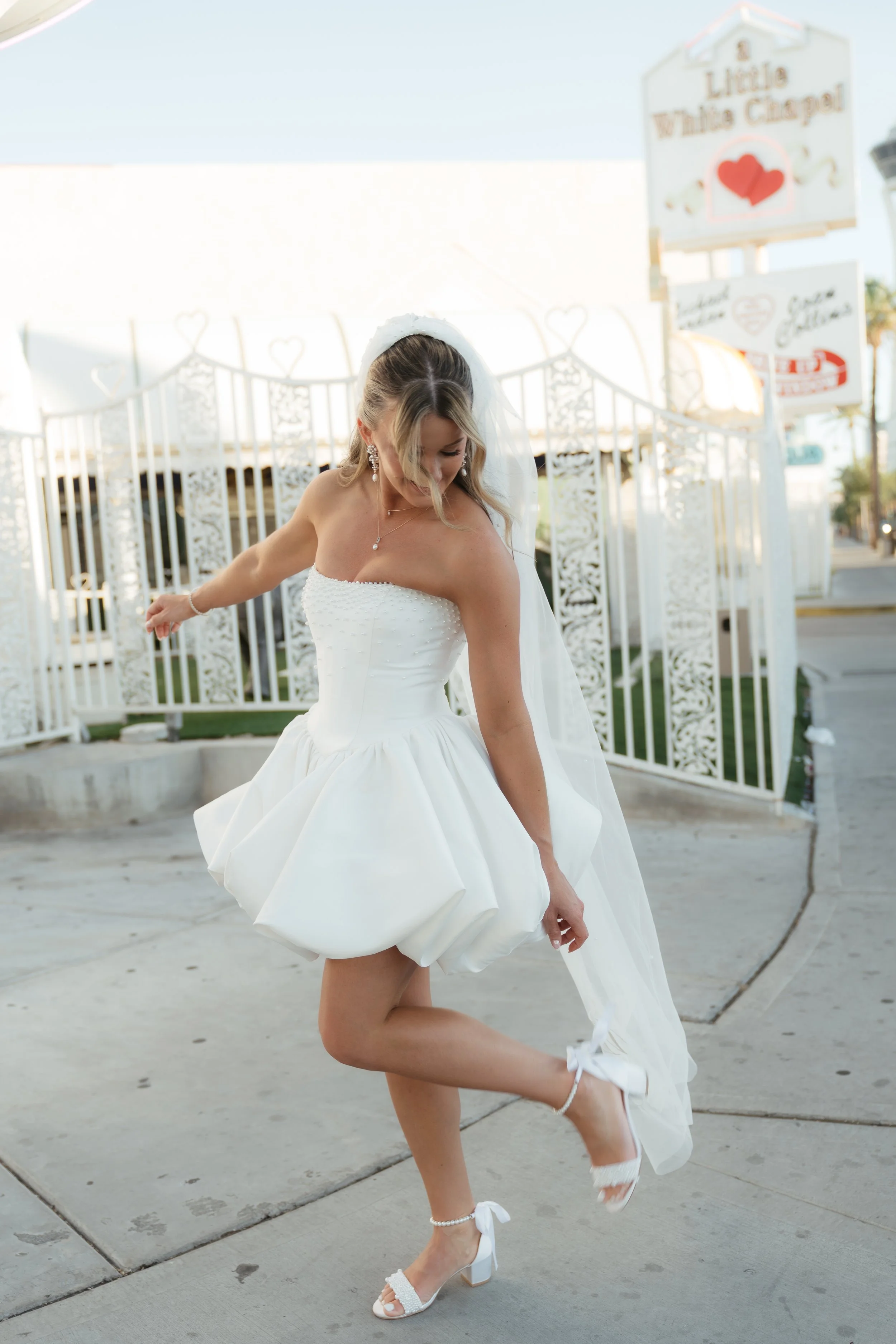 Bride in a white wedding dress spinning on a sidewalk outside, with a white decorative gate and signs in the background.