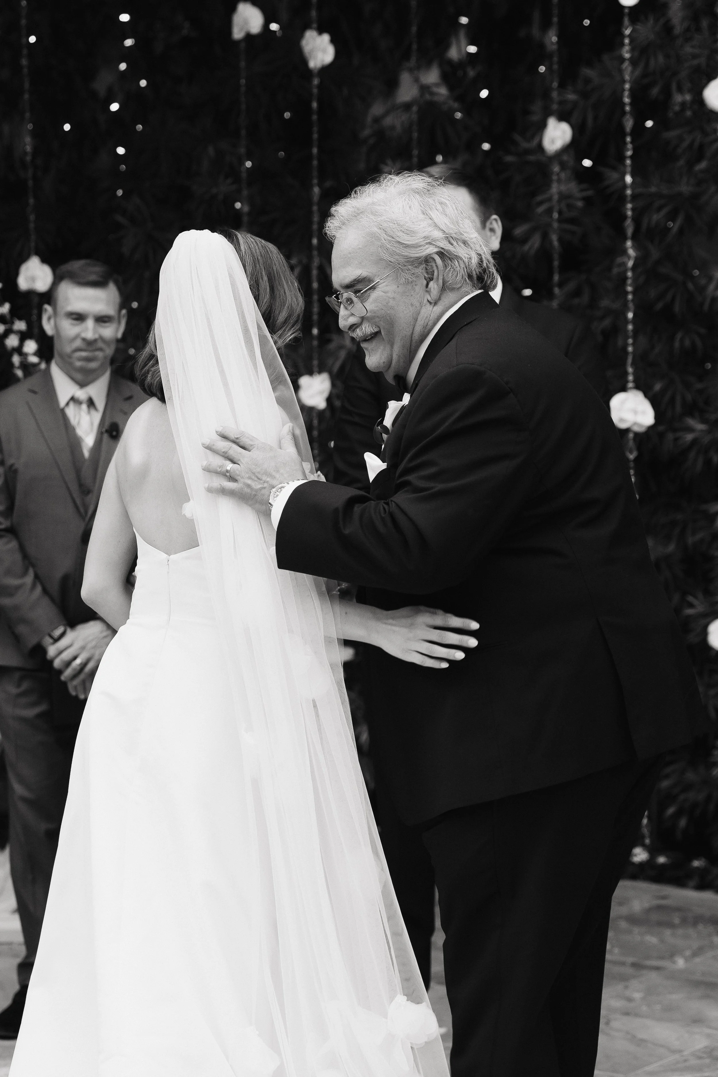 An elderly man in a tuxedo smiling as he places a wedding ring on a bride's finger during a wedding ceremony. The bride is wearing a white dress and veil. There is a man in a suit standing in the background. The scene is decorated with flowers and ha