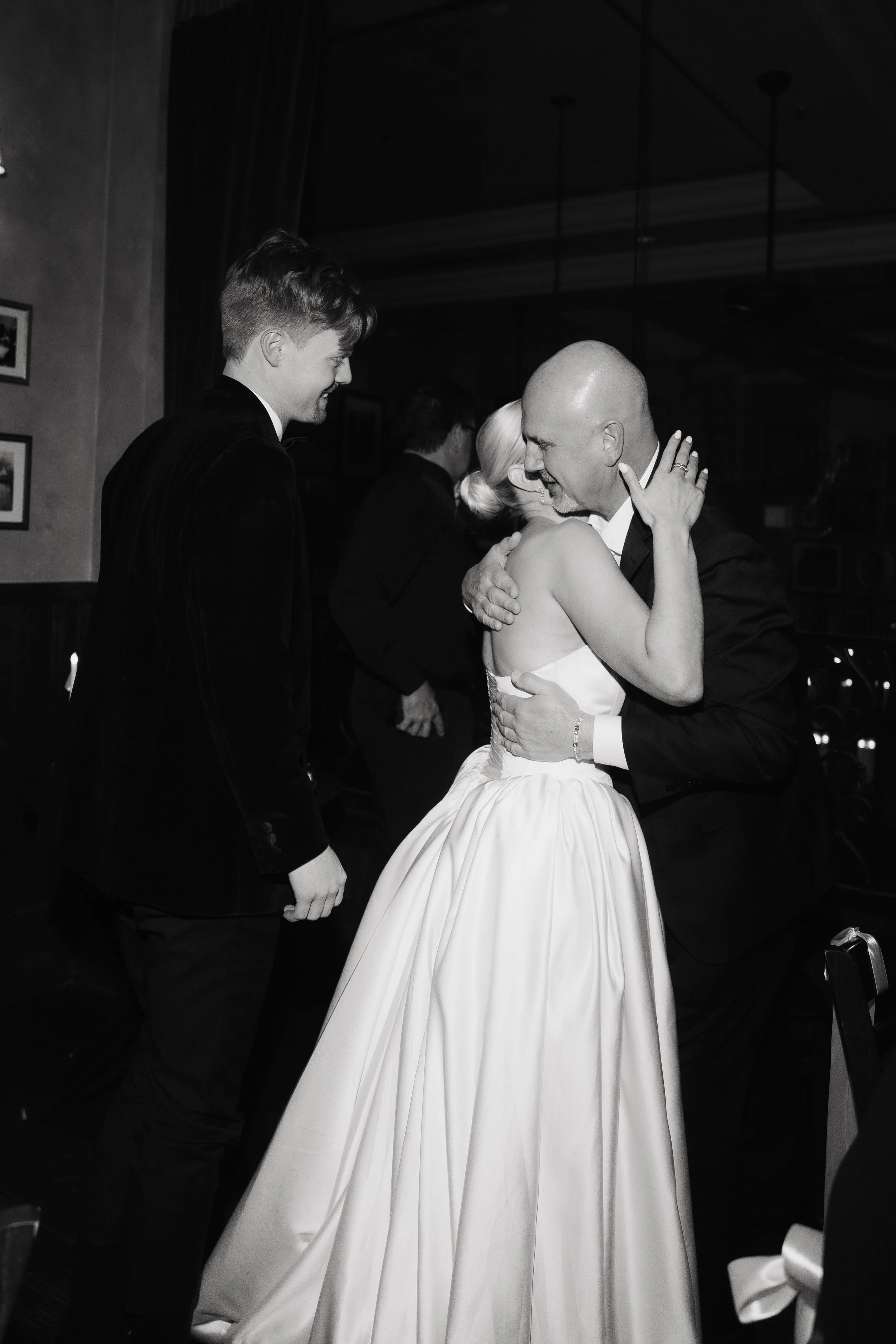 A bride and groom sharing a dance, with a man in a tuxedo standing nearby and smiling, in a black-and-white photo at a wedding reception.