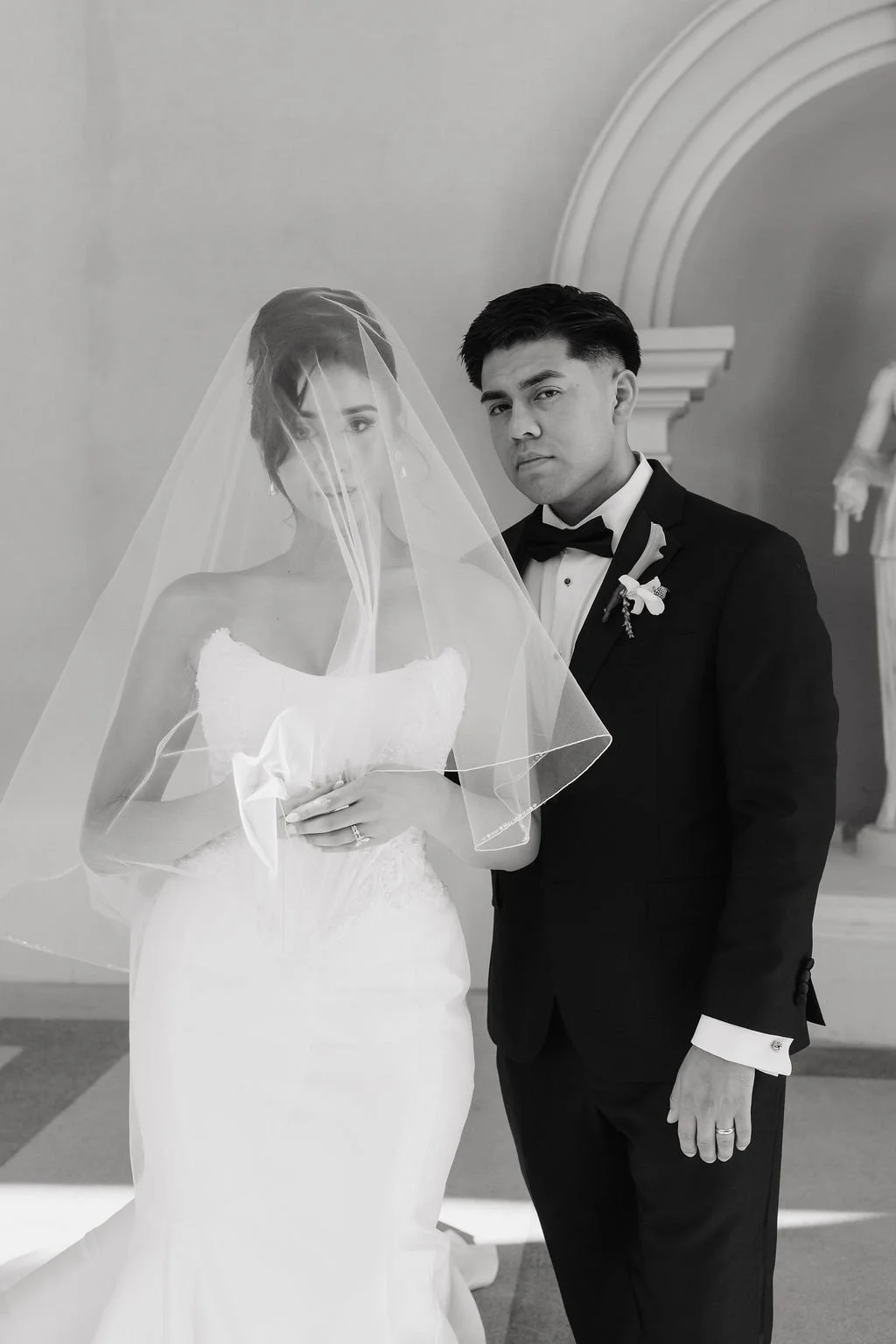 Black-and-white editorial portrait of a bride wearing a veil beside her groom in a tuxedo.
