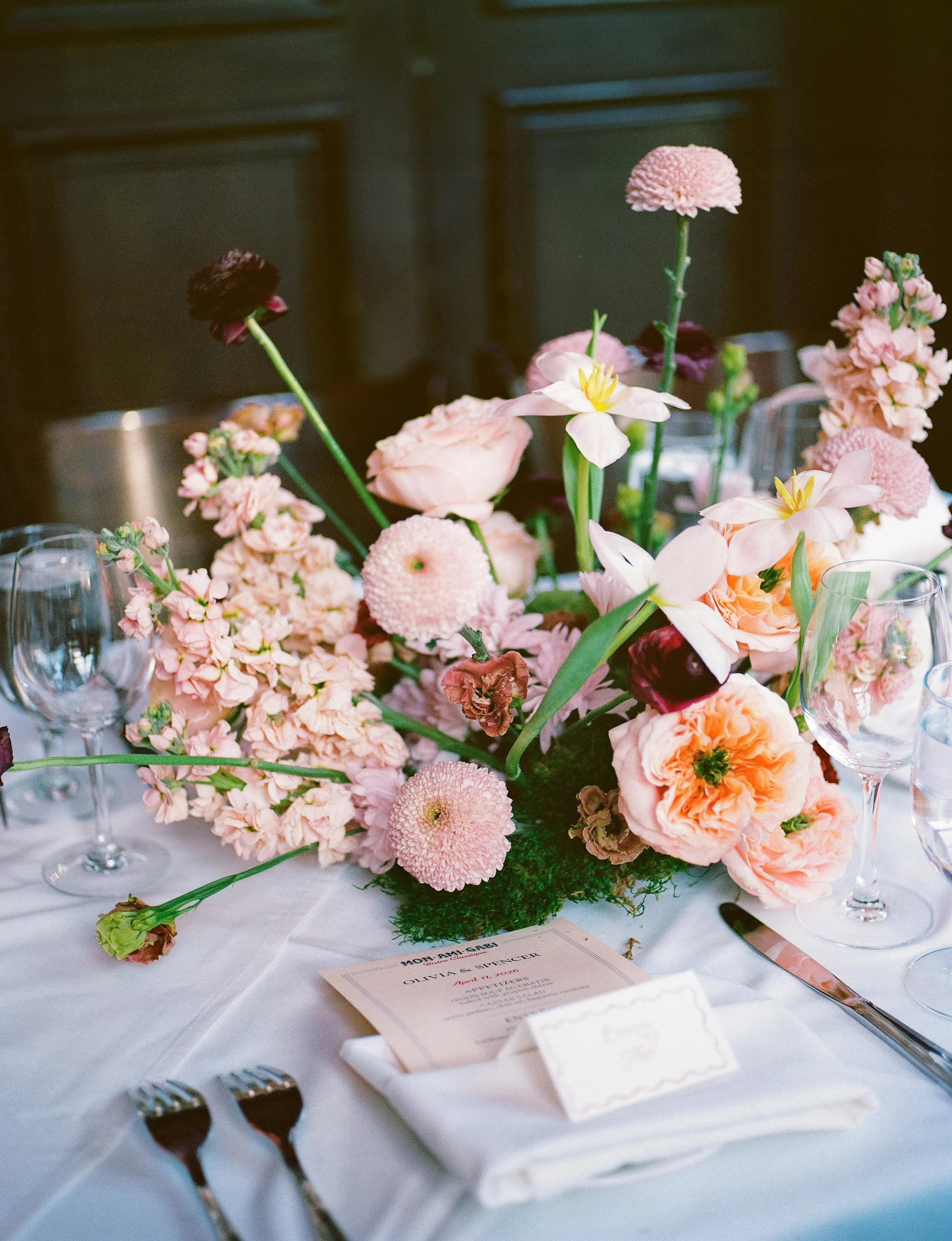 A floral centerpiece with pink and burgundy flowers on a table set for a formal event, with champagne flutes, silverware wrapped in a napkin, and a wedding invitation visible.