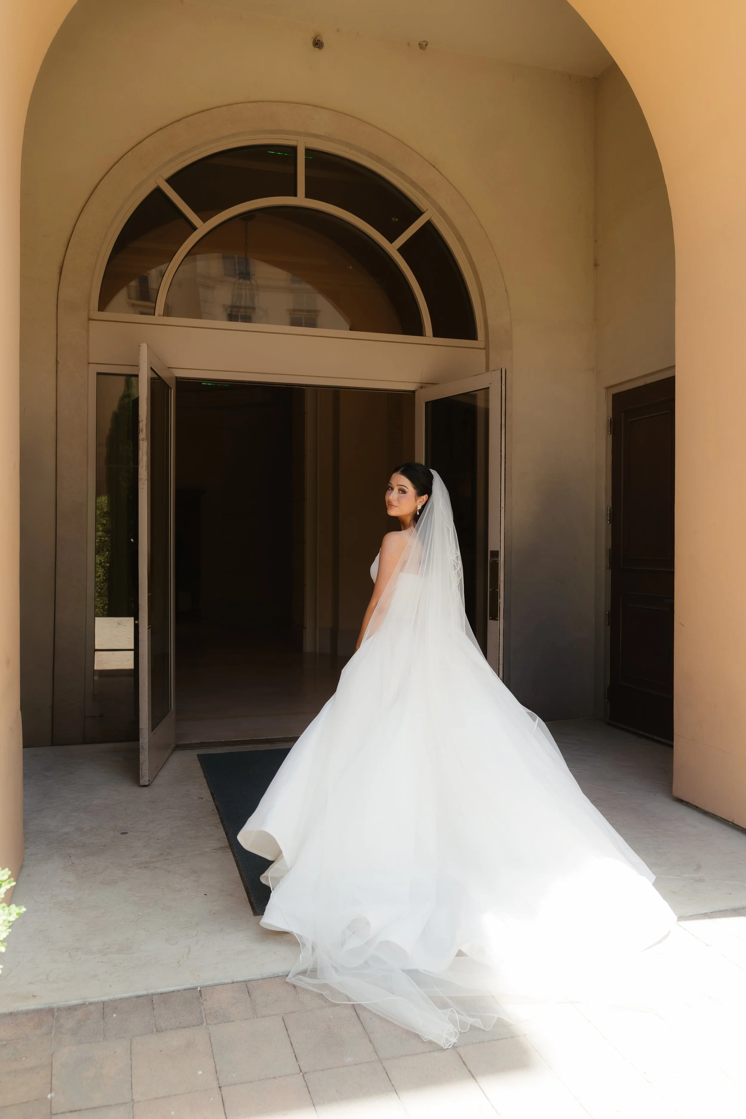 A bride in a white wedding gown with a long train stands in front of an open door with glass panels and an arched window above. The bride has dark hair styled in an updo and wears earrings and a veil. The setting appears to be outside a building with
