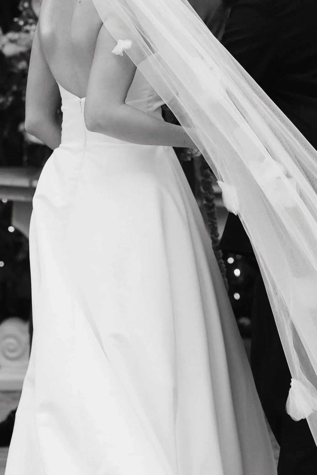 Black and white image of a bride in a wedding dress and a veil, standing next to a groom in a suit, focusing on their midsection.