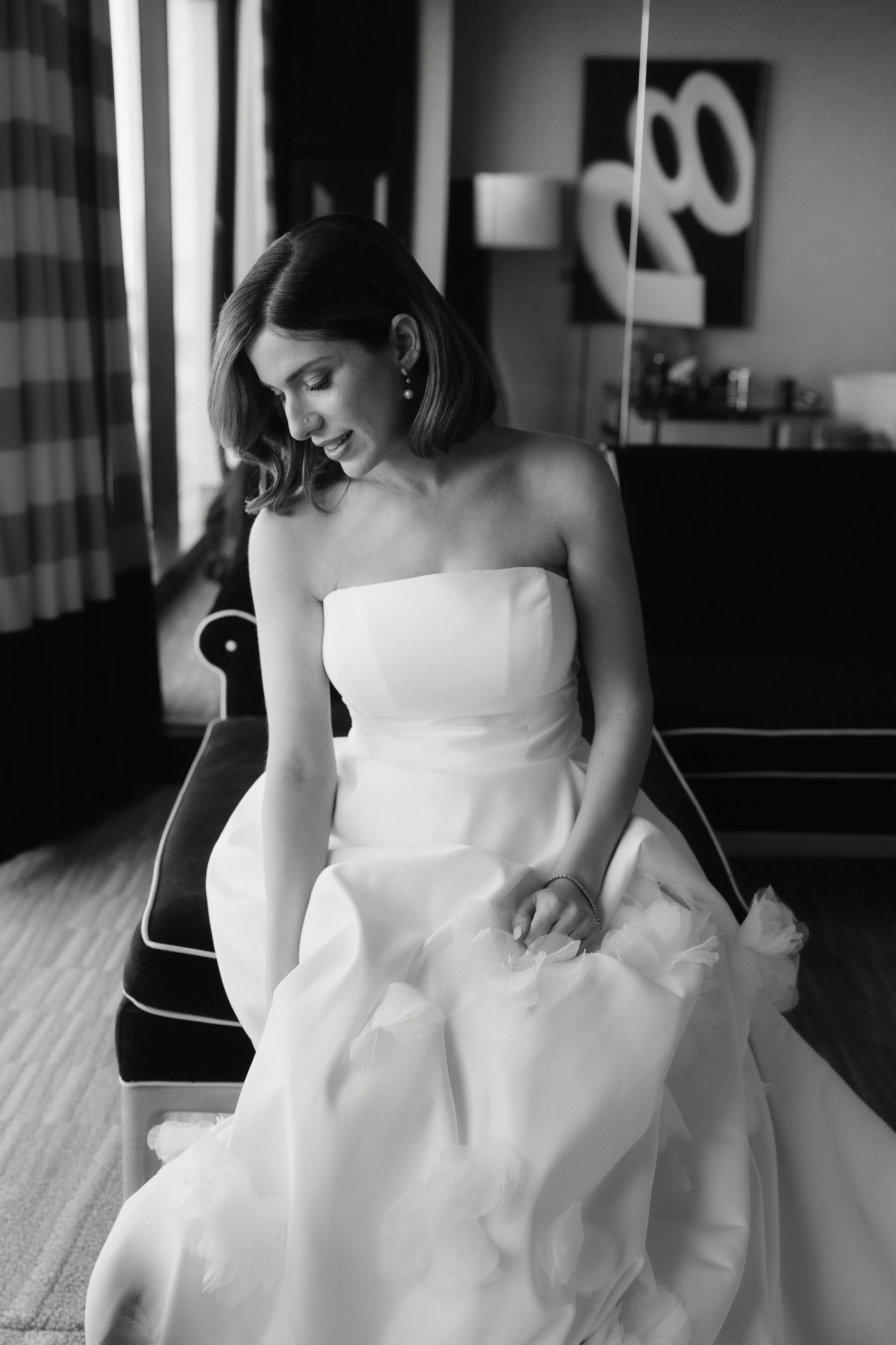 Black and white photo of a woman in a strapless wedding gown sitting on a bench, looking down, with a modern decor background.