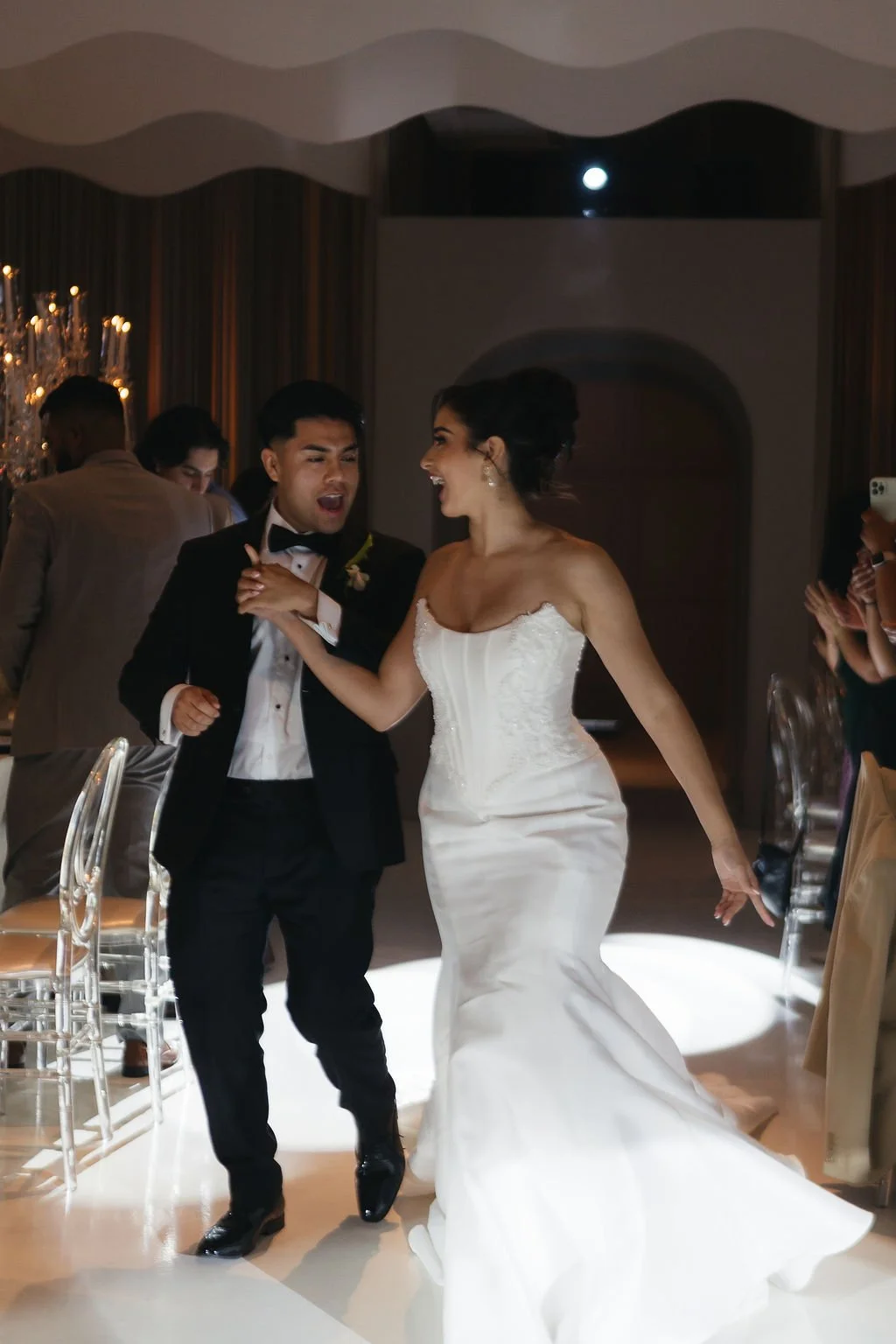 Newlyweds entering their wedding reception hand in hand as their guests welcome them.
