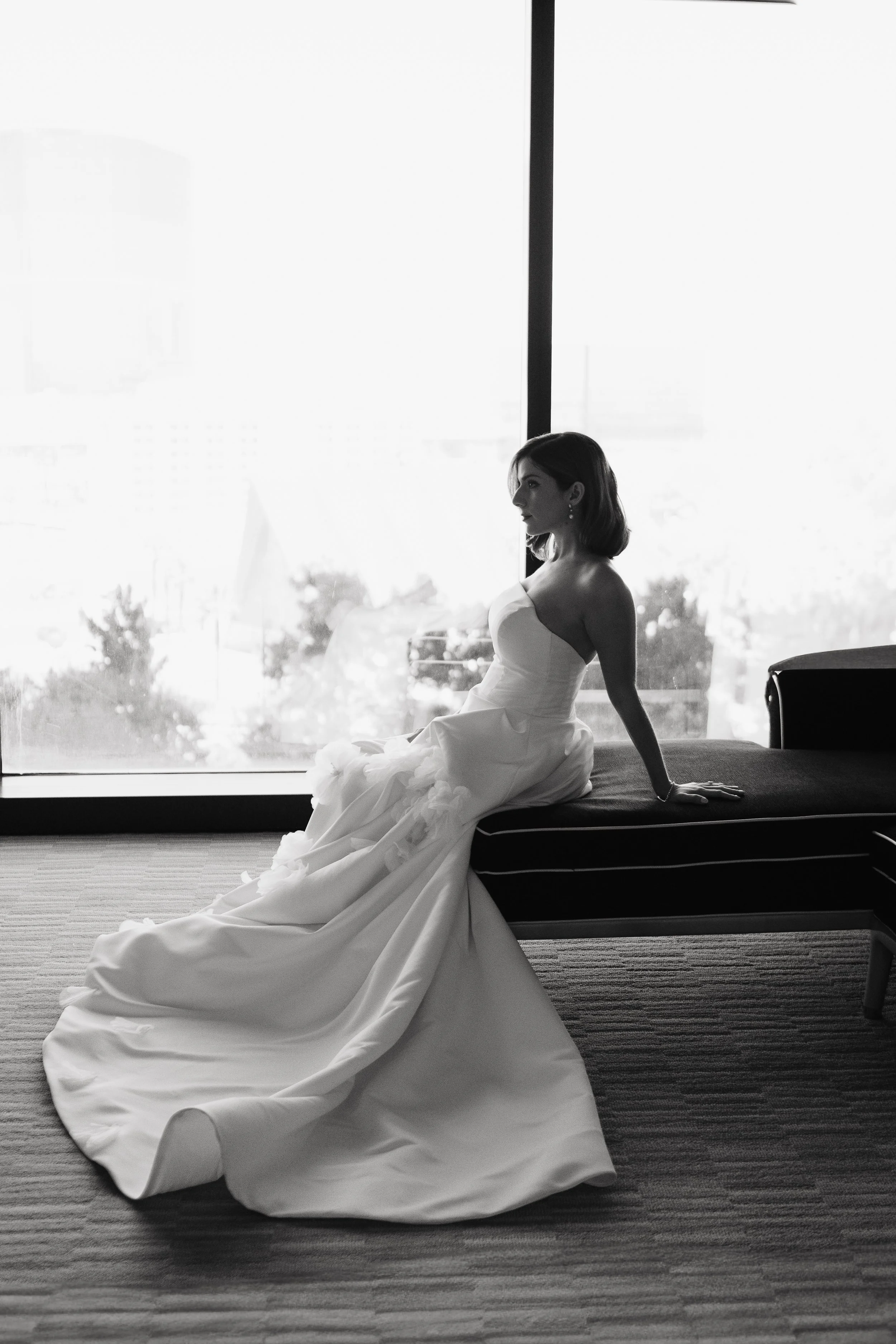 A woman in a wedding dress sitting on a bench by a large window, looking to her left with a calm expression. The cityscape can be seen outside the window.
