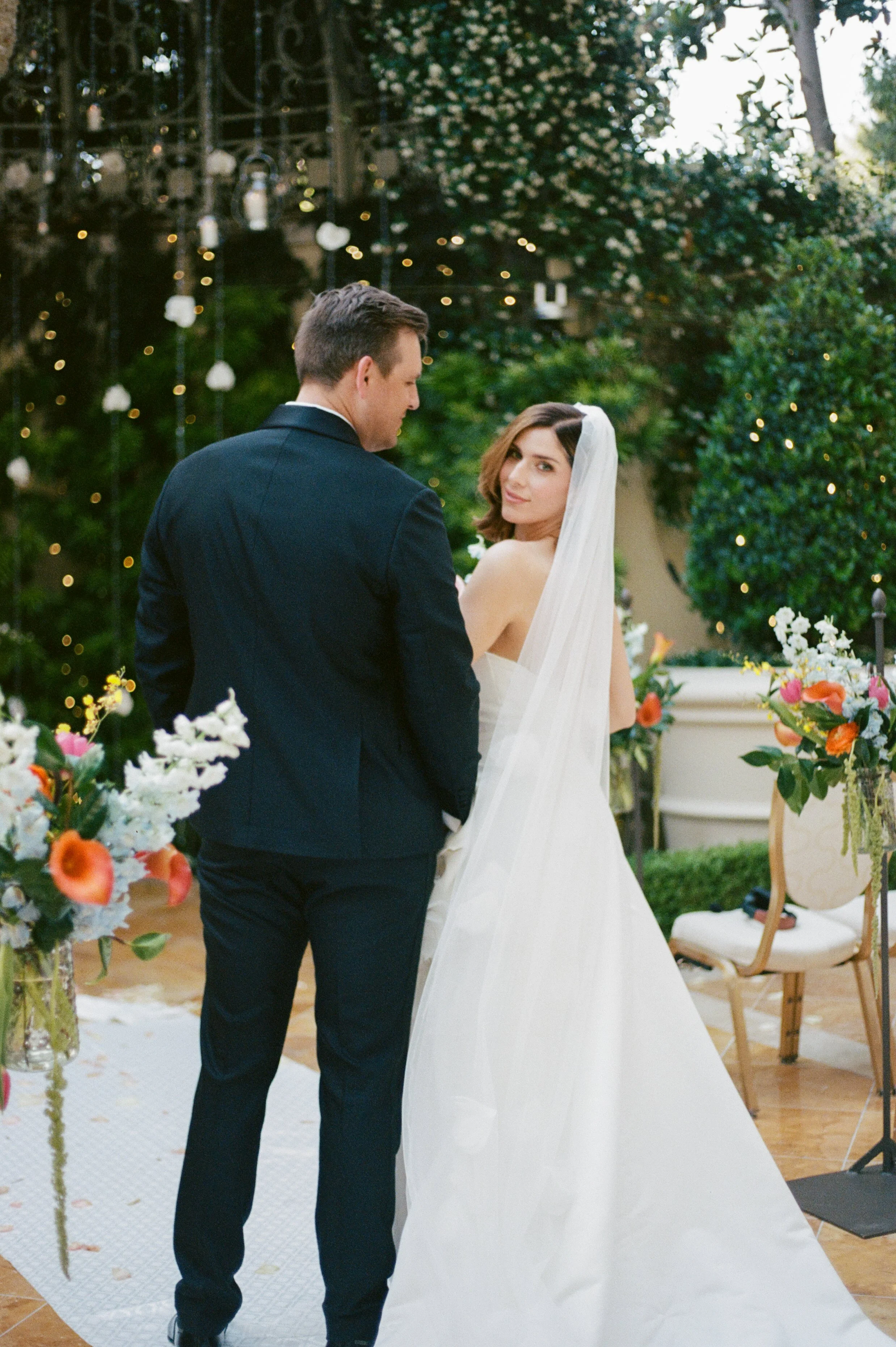 A groom and bride sharing a moment during their wedding ceremony outdoors, surrounded by flowers and greenery.