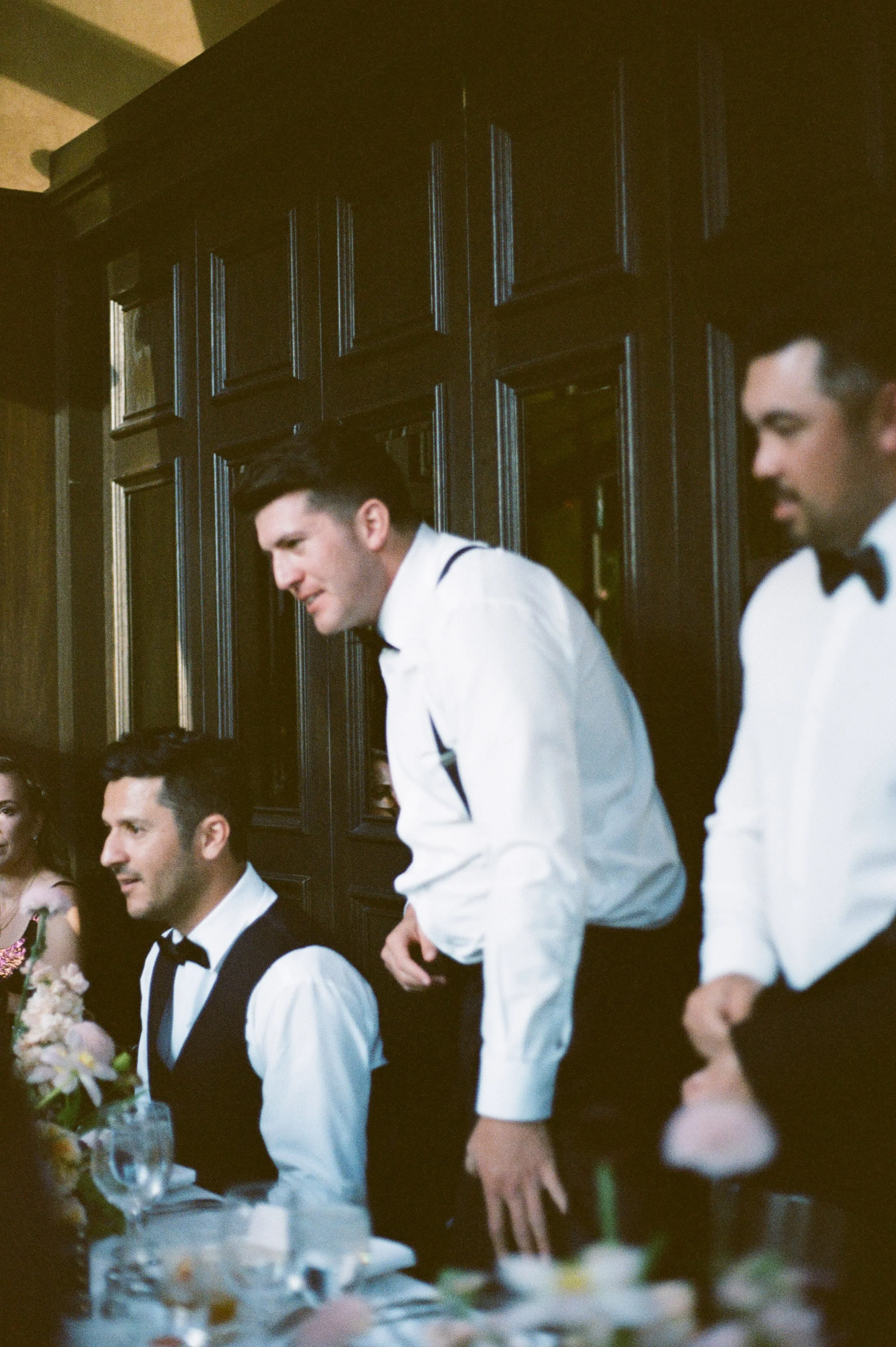 Two men dressed in white shirts, black bow ties, and black vests sitting at a Vegas wedding reception