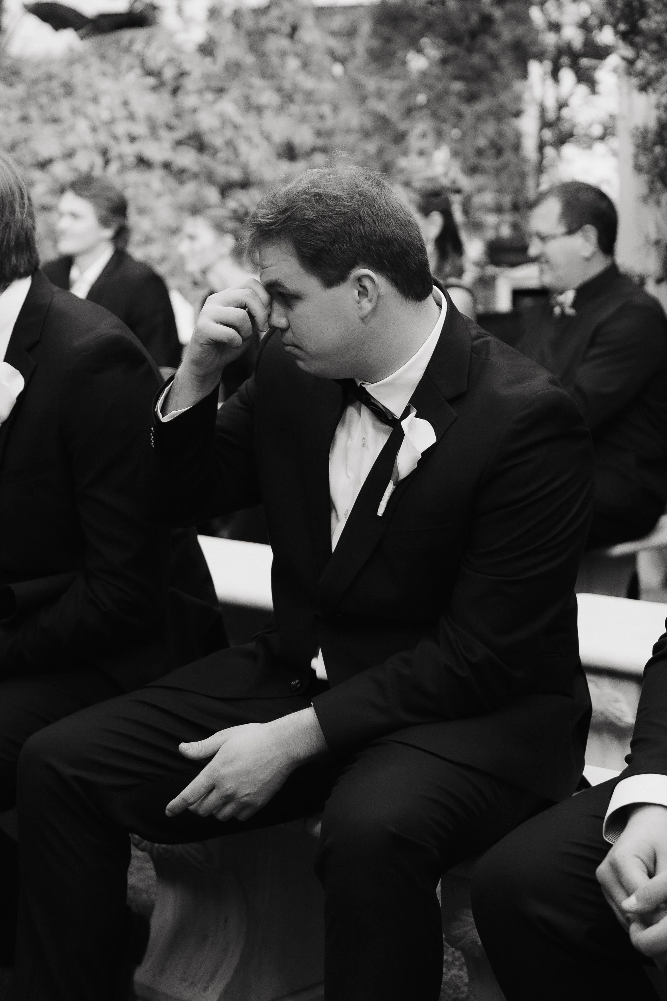 Black and white photo of a group of people dressed in formal attire, sitting outdoors, with trees and rocks in the background. One man in the center has his hand on his face, appearing deep in thought.