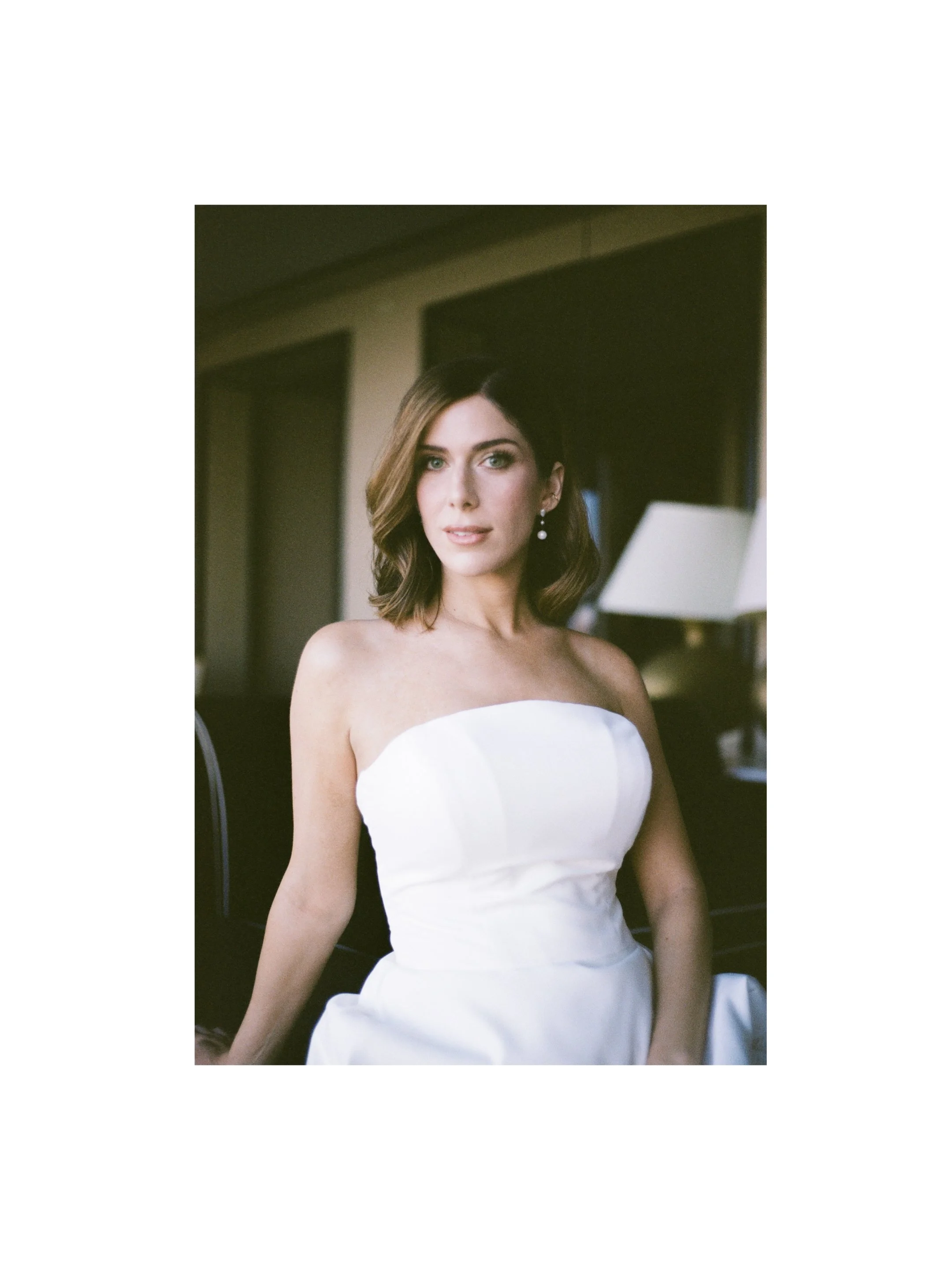 A woman with shoulder-length brown hair and blue eyes wearing pearl earrings and a strapless white dress, sitting indoors in front of a dark background with a lamp and window.