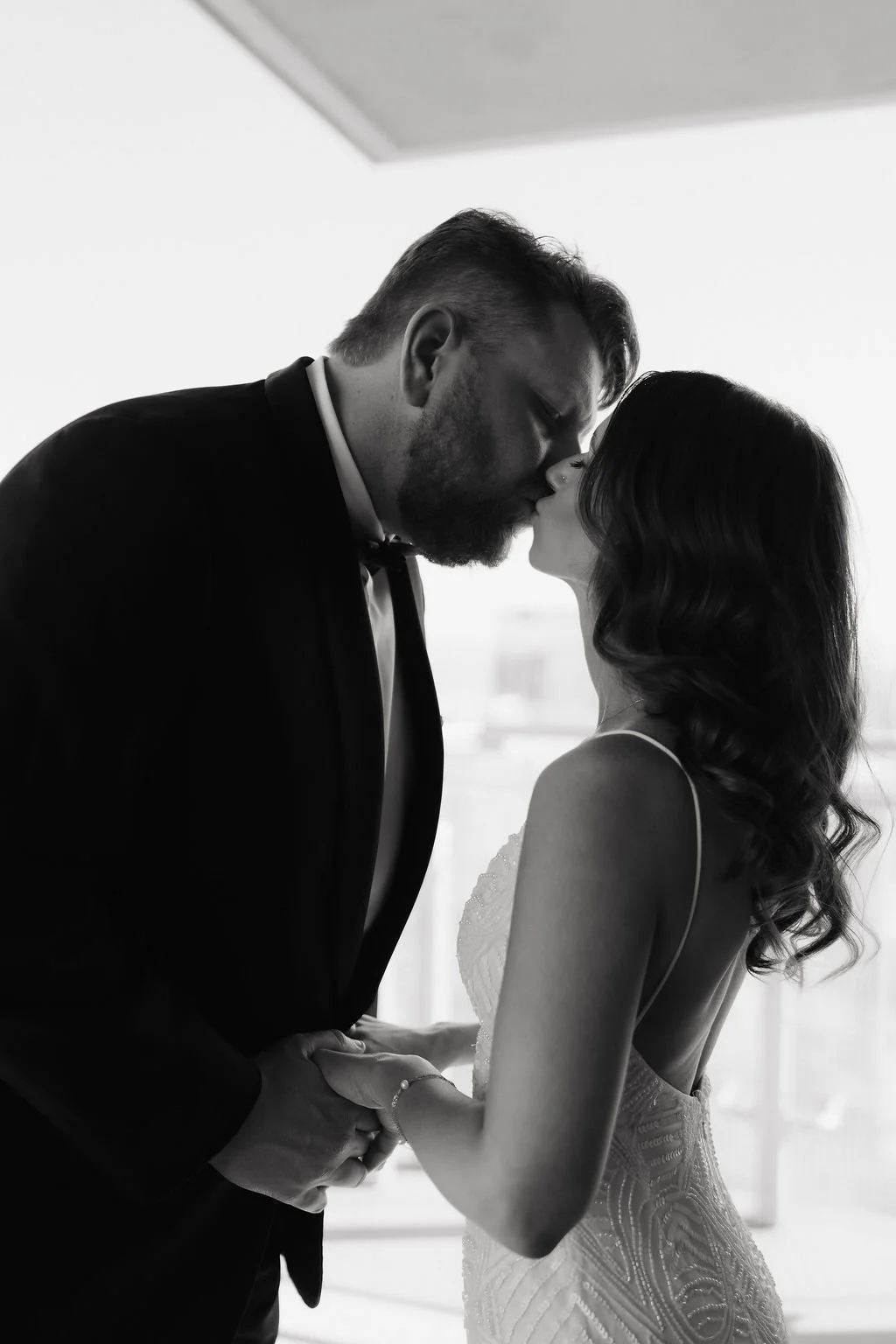 A black-and-white photo of a couple sharing a kiss, dressed in formal wedding attire, indoors with natural light.