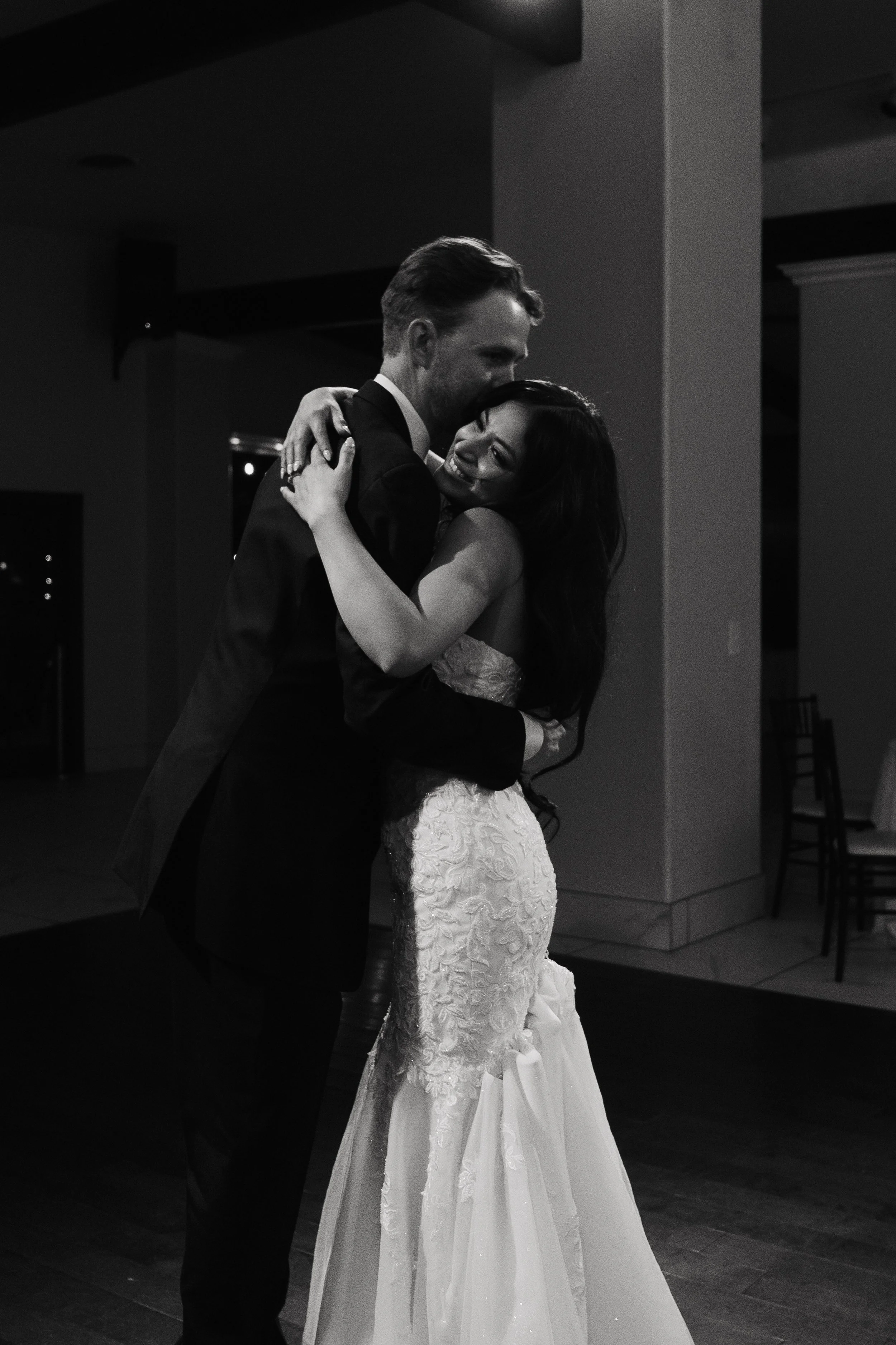 A black and white photo of a bride and groom dancing closely at their wedding reception indoors, with the groom gently embracing the bride.