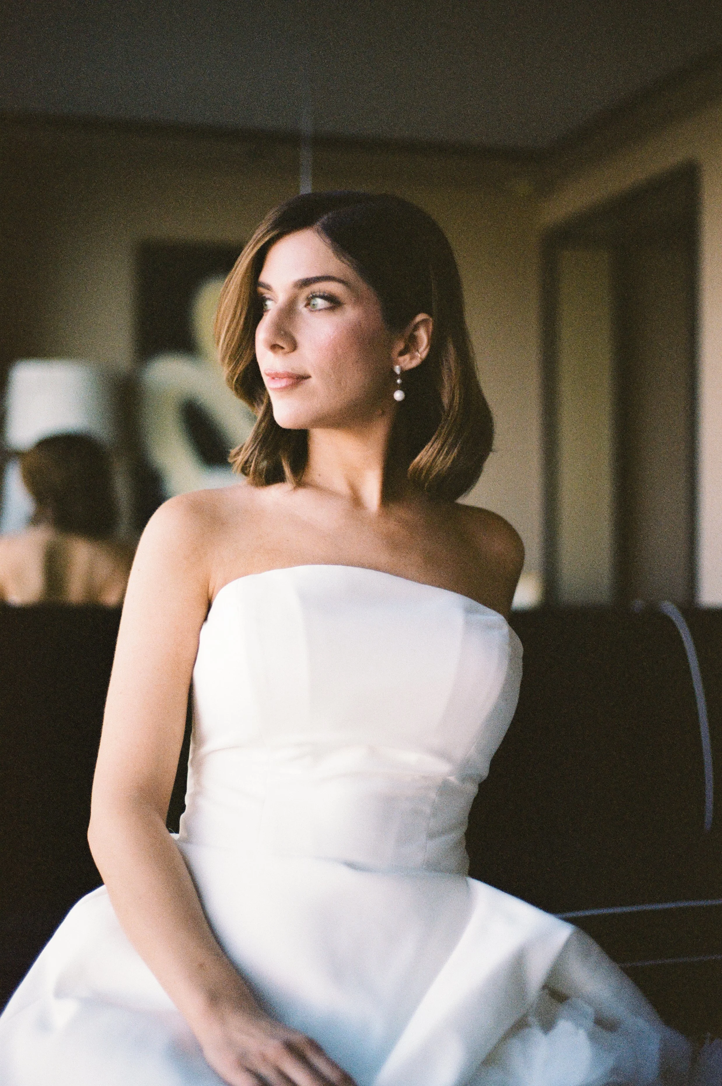 A woman with shoulder-length brown hair wearing a strapless white dress and pearl earrings, looking to her left in a dimly lit room.