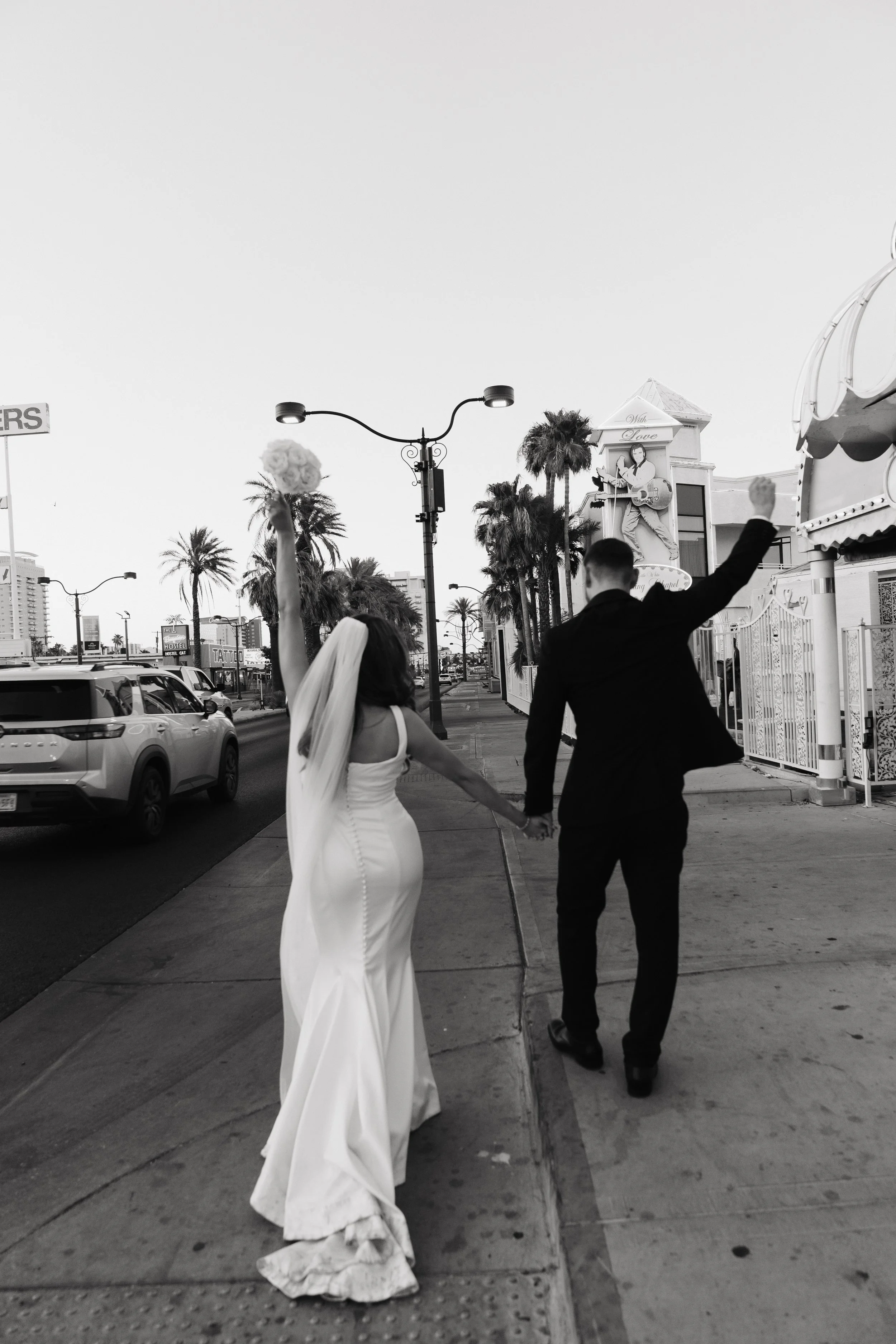 A bride and groom holding hands as they walk along a city sidewalk with palm trees in the background, celebrating their wedding.