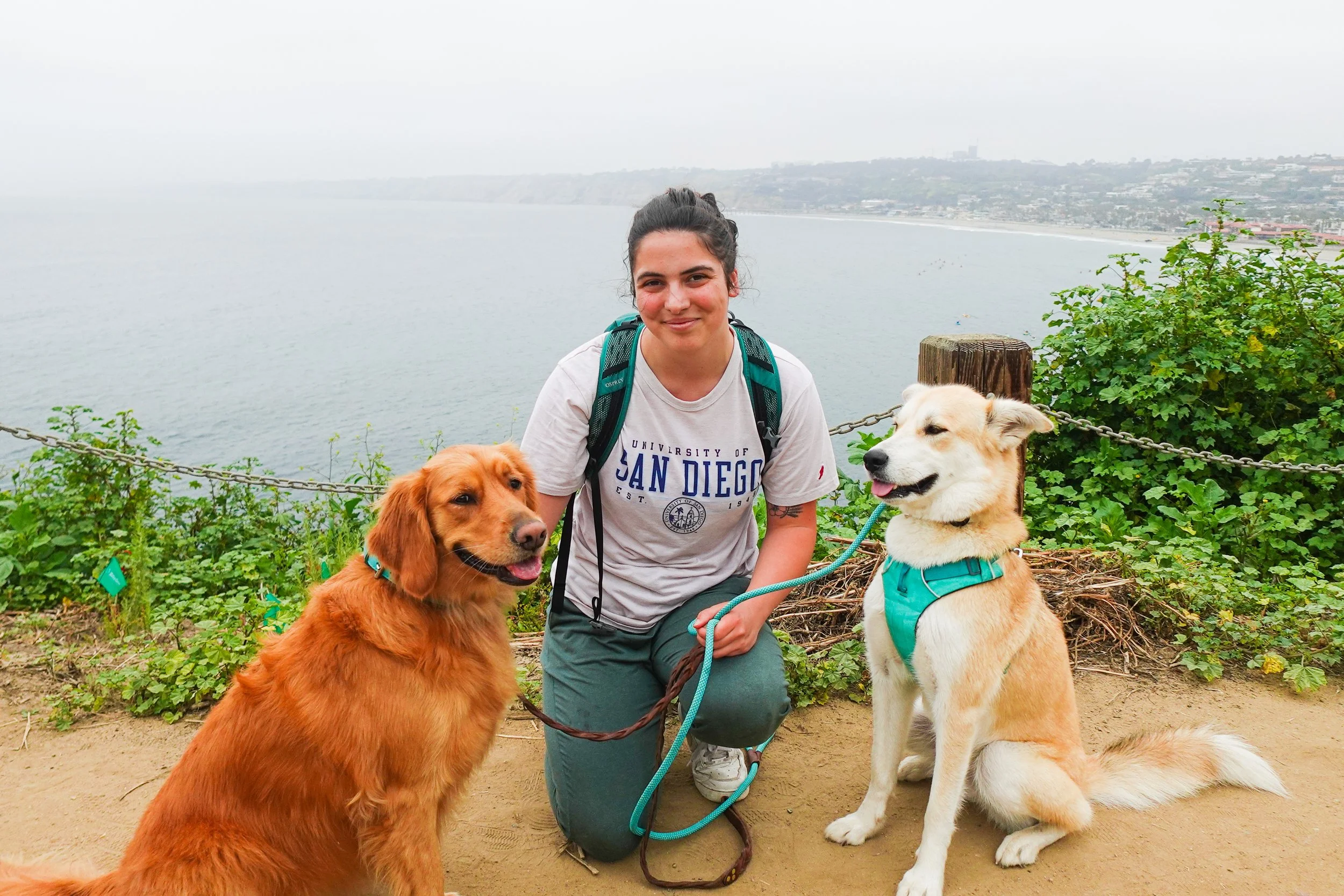 A smiling woman with dark hair tied back, wearing a white T-shirt and jeans, kneeling on a dirt path beside two dogs, with a foggy coastline and water in the background.