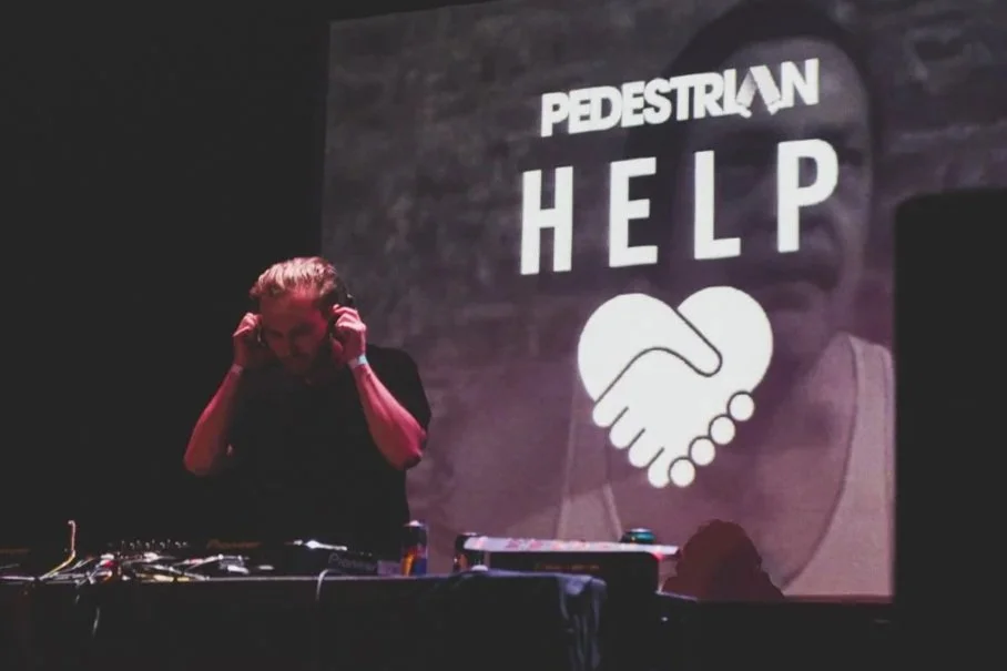 DJ or performer adjusting headphones in front of a large screen displaying the logo and message for Pedestrian Help, featuring a handshake icon.