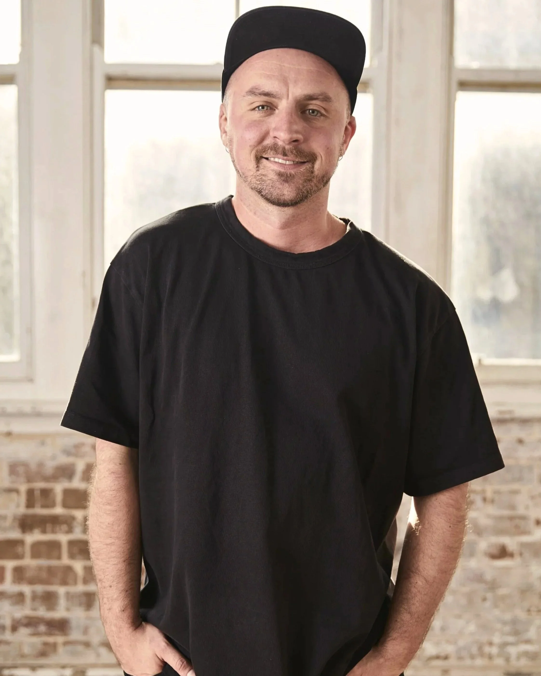James McManus with a black baseball cap, wearing a black t-shirt, standing indoors in front of large windows with a brick wall below.