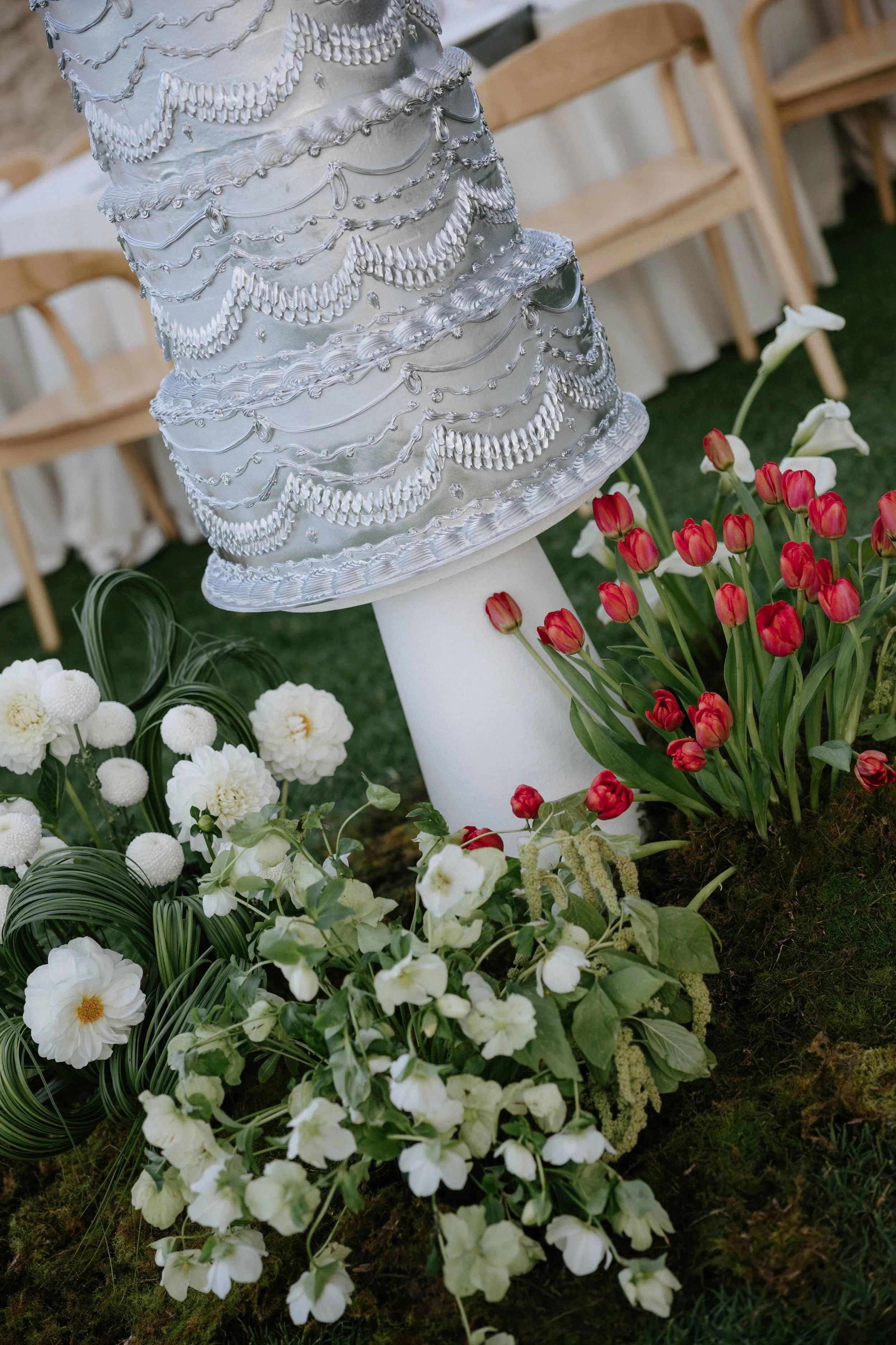 A tiered wedding cake decorated with silver piping and icing, surrounded by various white and red flowers, in an outdoor setting with wooden chairs in the background.