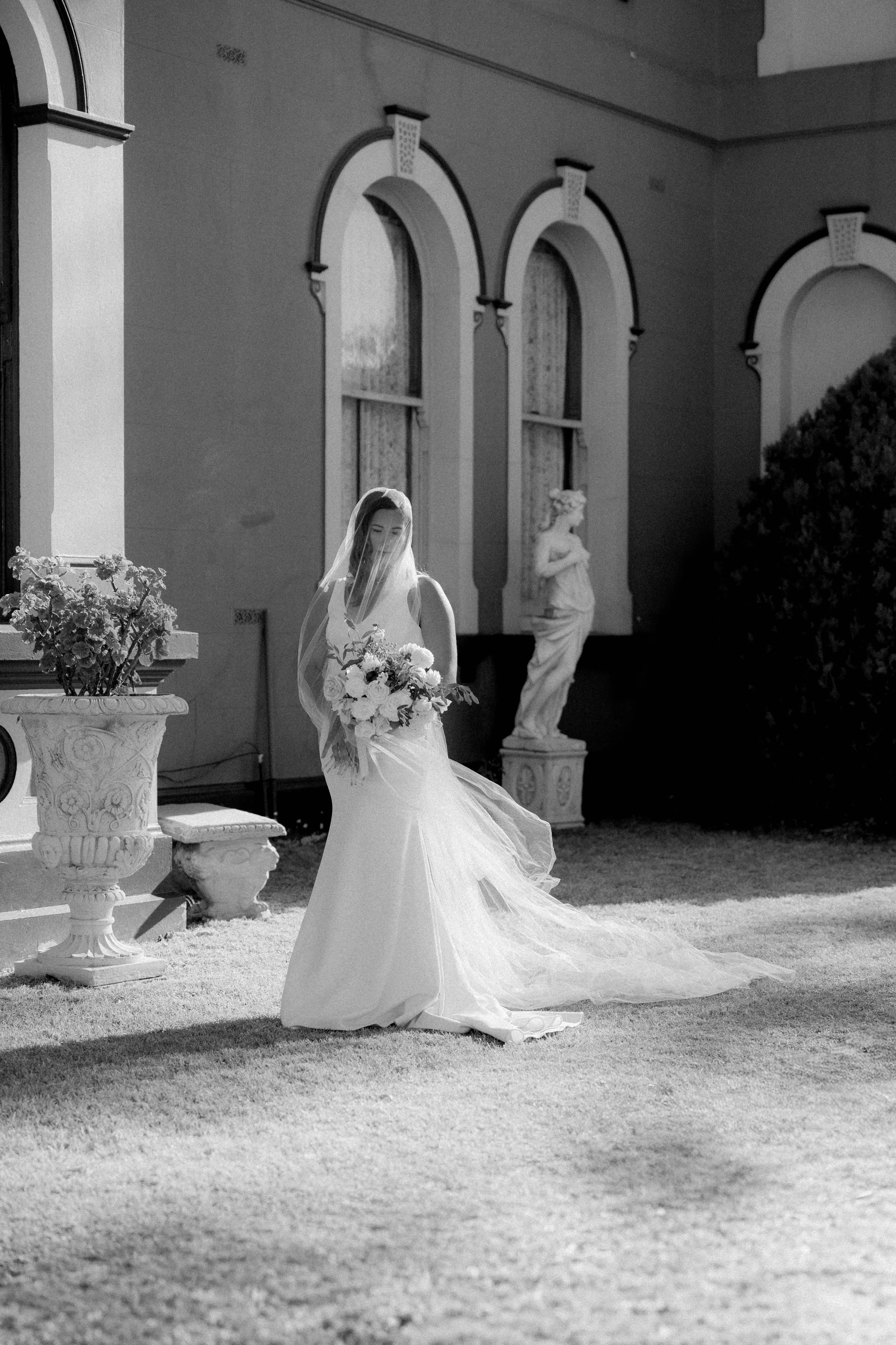 A woman in a wedding dress holding a bouquet of flowers, standing outdoors in front of a building with large windows and classical sculptures, in black and white.