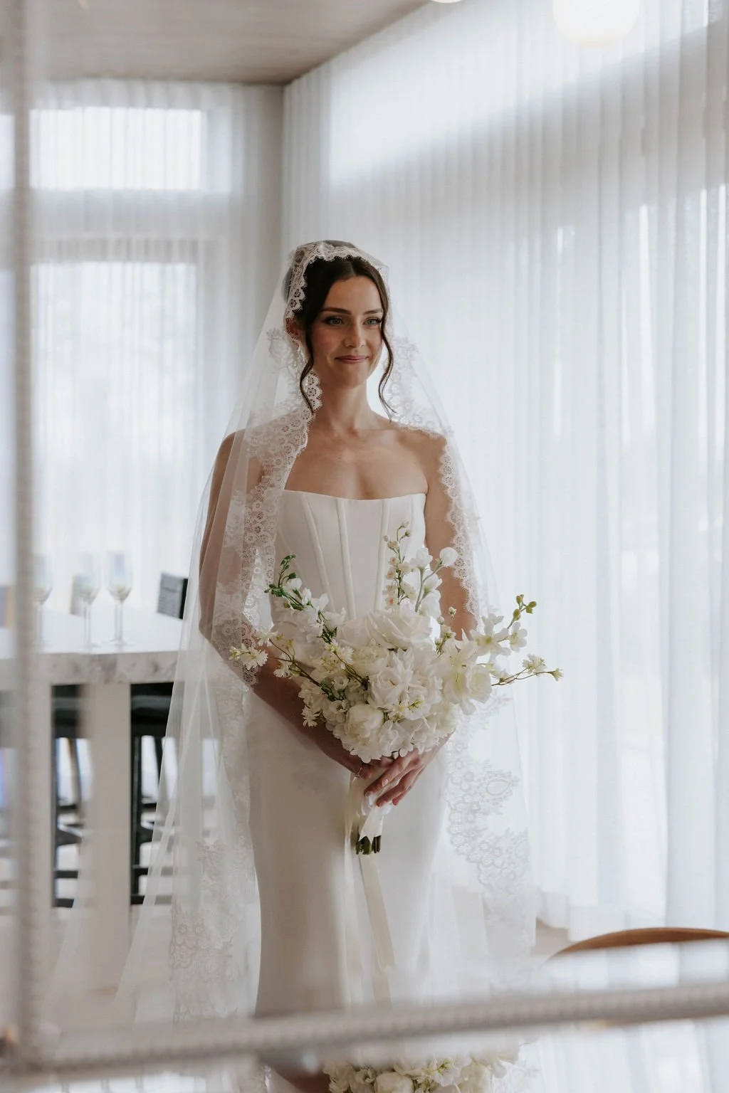 A bride stands indoors with white curtains and natural light, wearing a strapless white wedding dress, lace veil, and holding a large bouquet of white flowers.