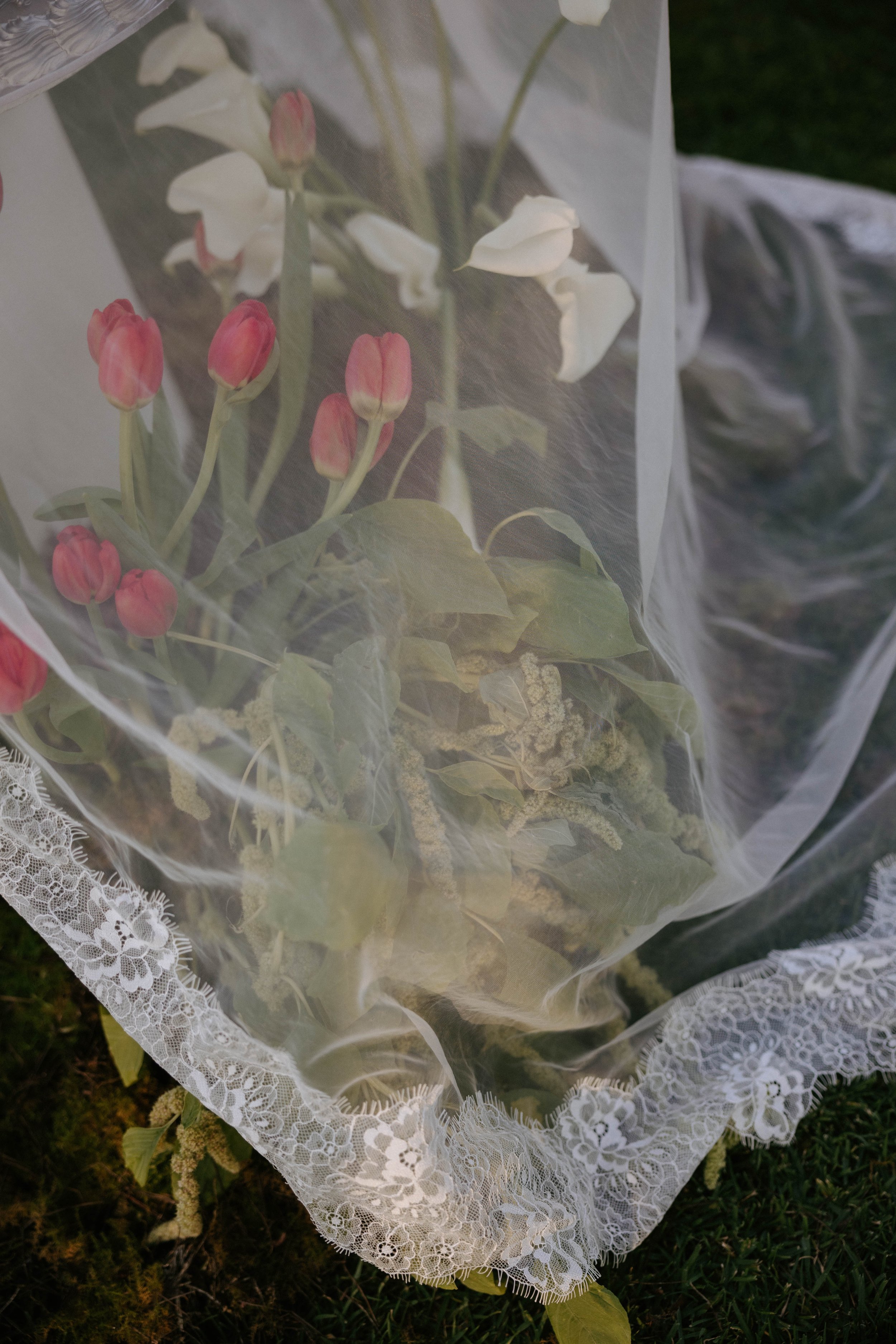 A bouquet of pink and white flowers wrapped in sheer lace and plastic, resting on grass.