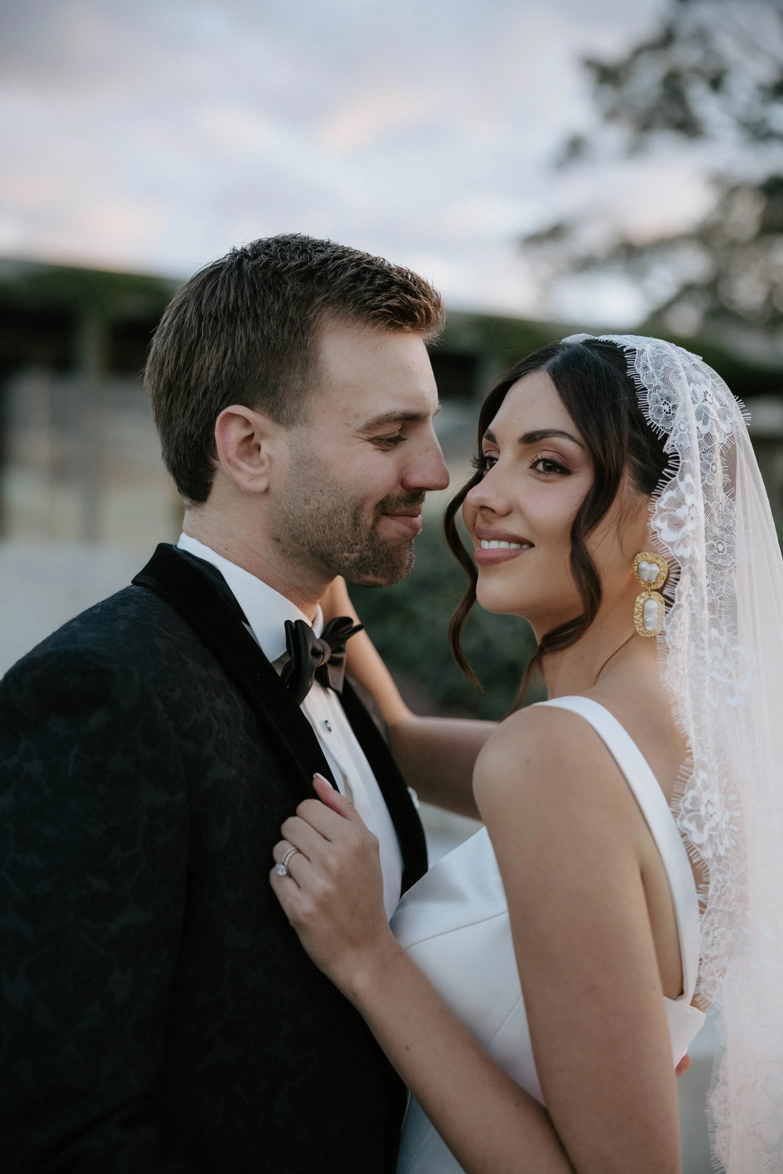A wedding couple, the groom in a black tuxedo and bow tie, and the bride in a white dress with a lace veil, looking at each other and smiling, outdoors during sunset.