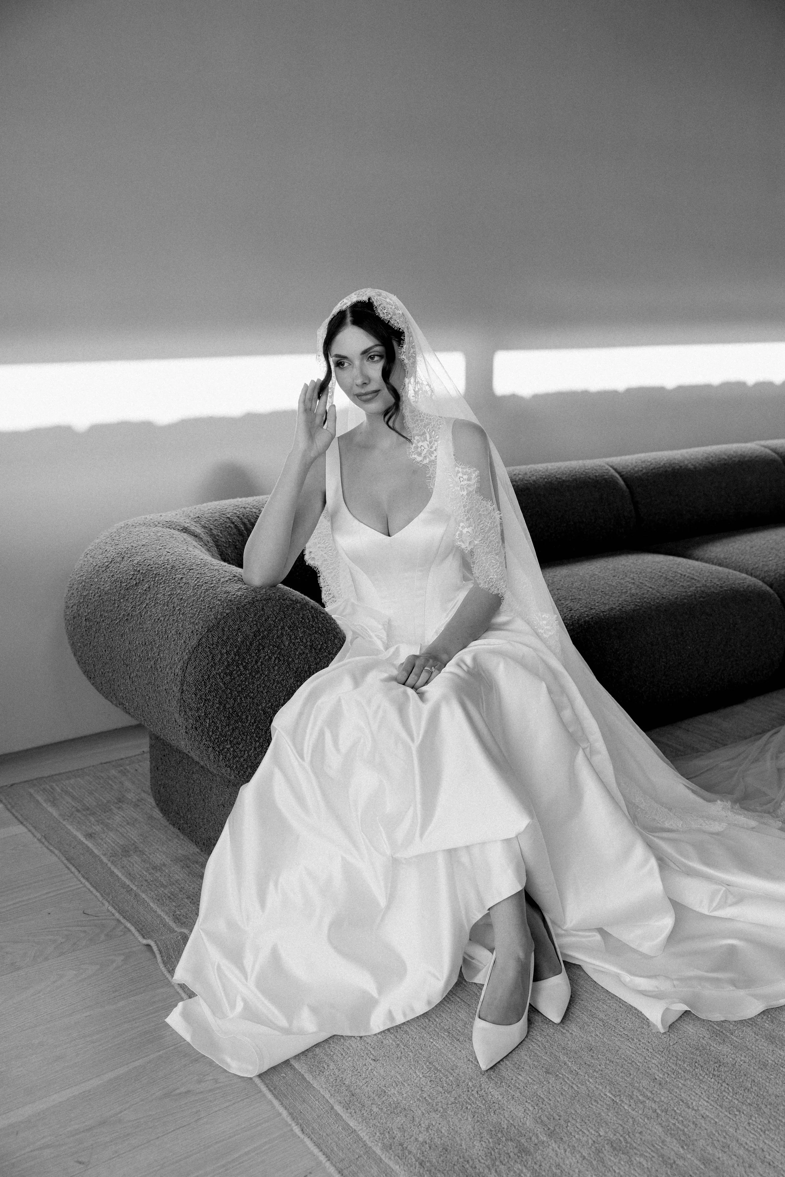 Black and white photo of a woman in a wedding dress sitting on a curved sofa in a room with hardwood floors and a large window. She is wearing a veil and high-heeled shoes, with her right hand near her face and her left hand resting on her lap.