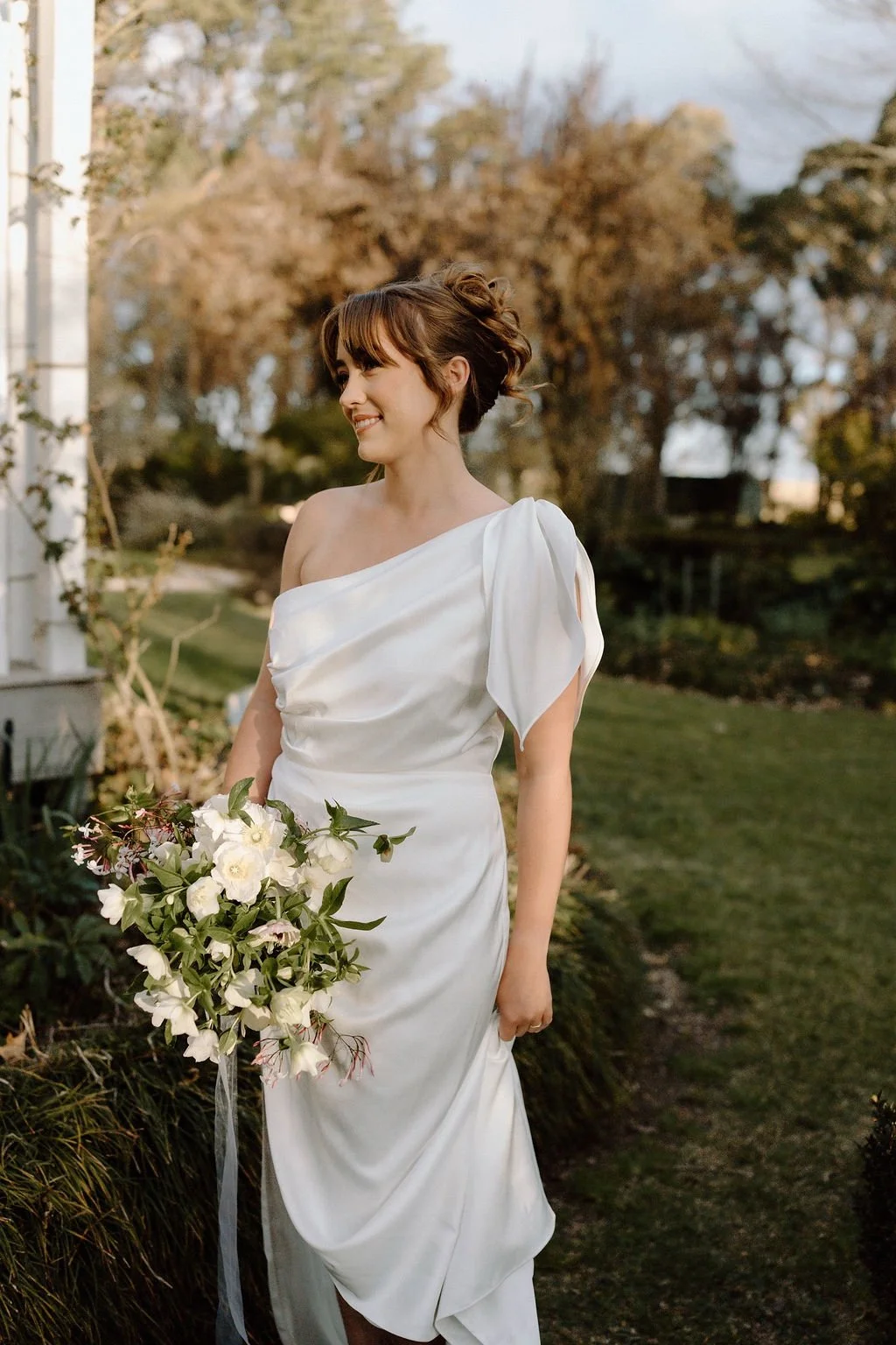 A bride in a white gown holding a bouquet of white flowers, standing outdoors in a garden.