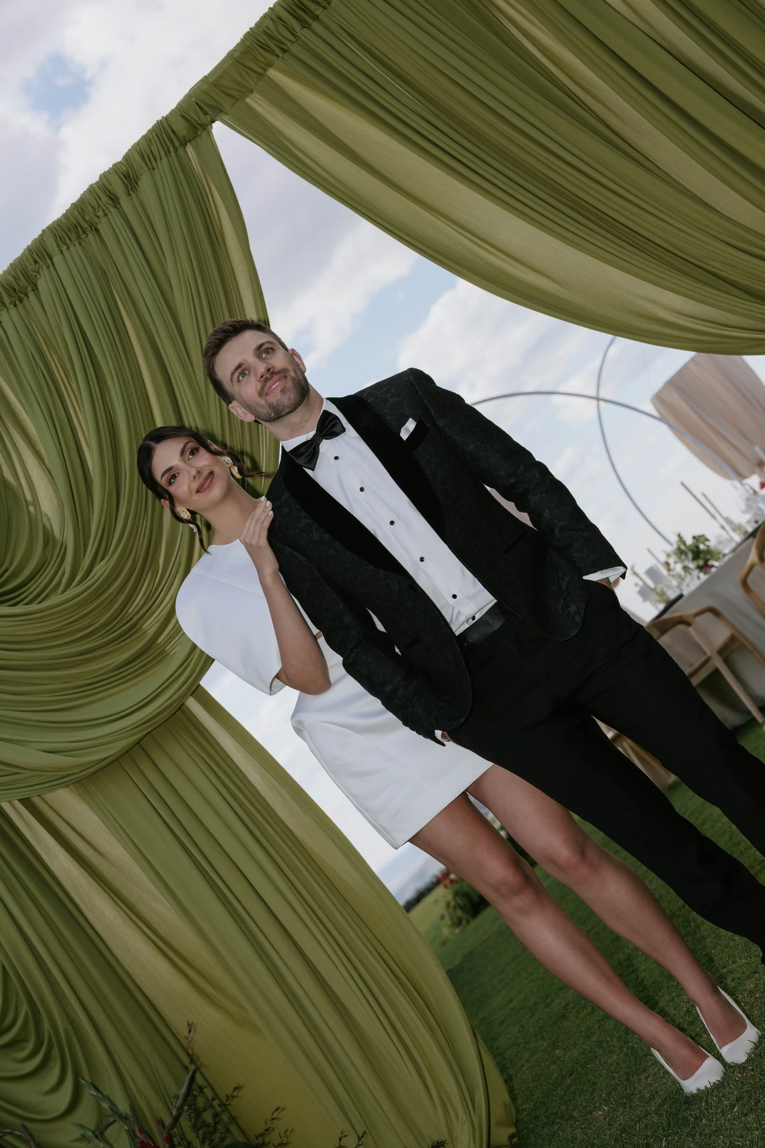 A man and a woman dressed in formal attire standing under a green fabric canopy outdoors, with a cloudy sky and some greenery in the background.