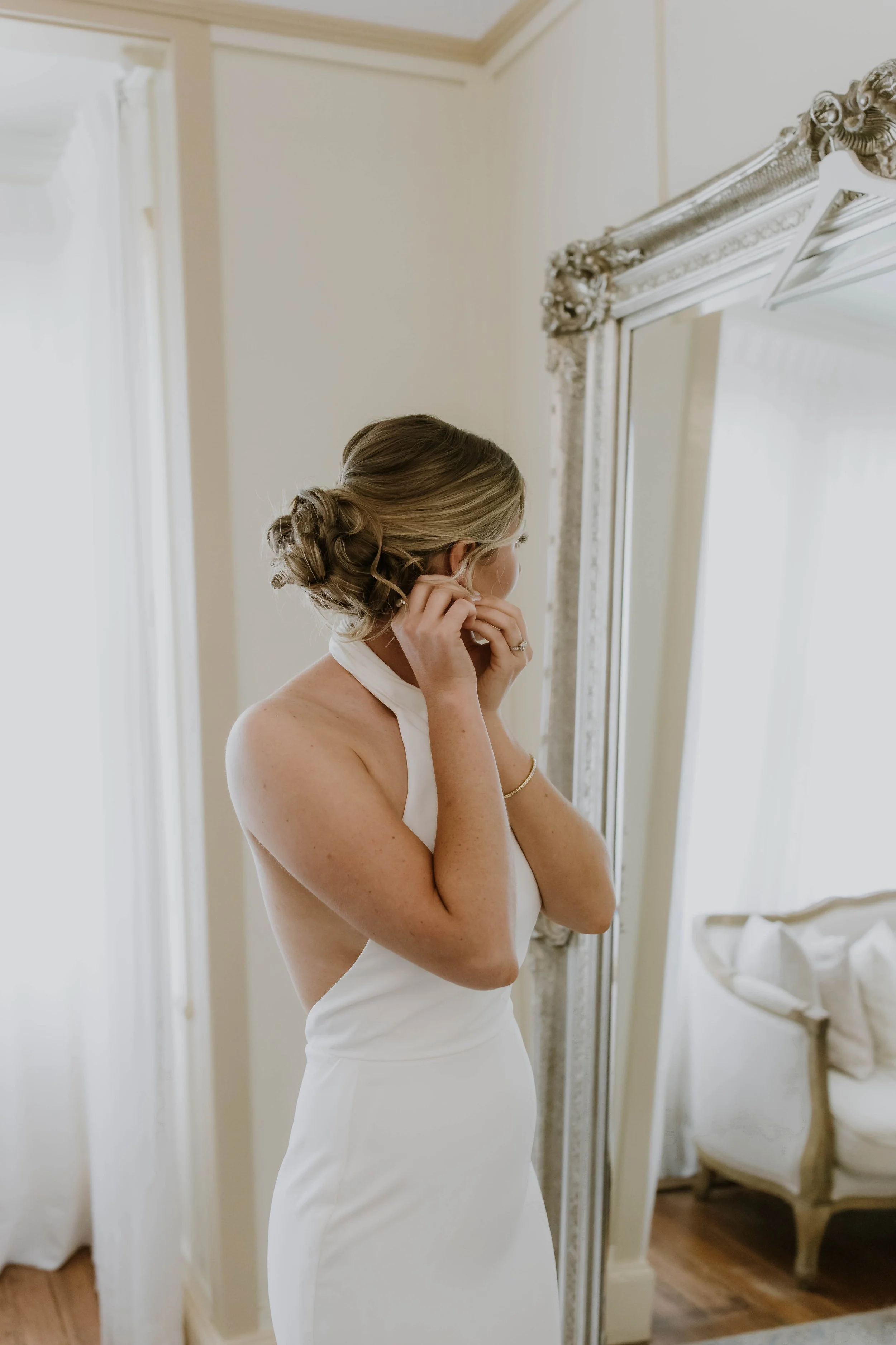 A woman with styled hair putting on earrings, dressed in a white dress, standing in front of a large mirror in a room with cream-colored walls and classic furniture.