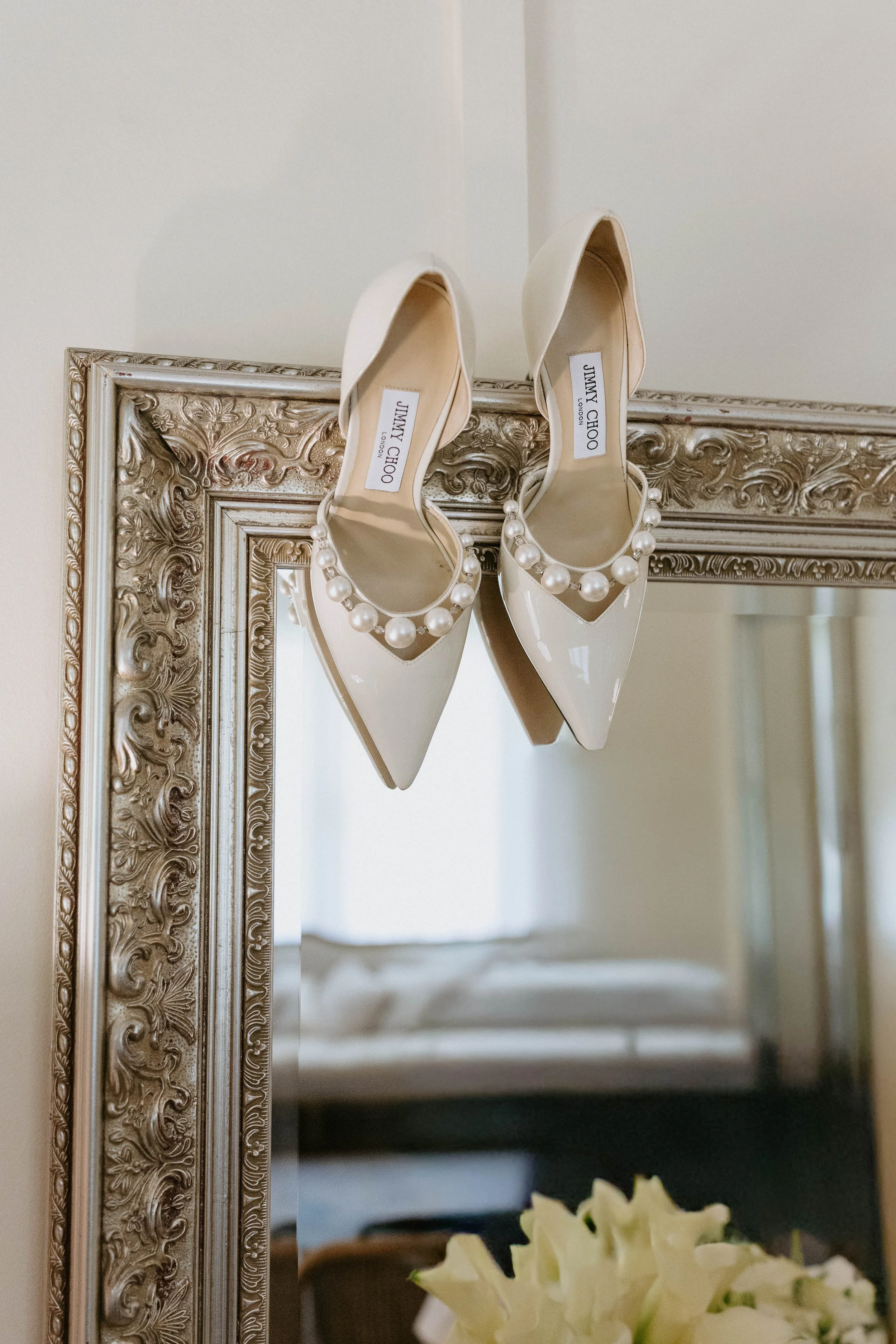 A pair of white Jimmy Choo high heels with pearl embellishments, hanging upside down in front of a decorative mirror with an ornate silver frame, with a bouquet of white flowers visible at the bottom.