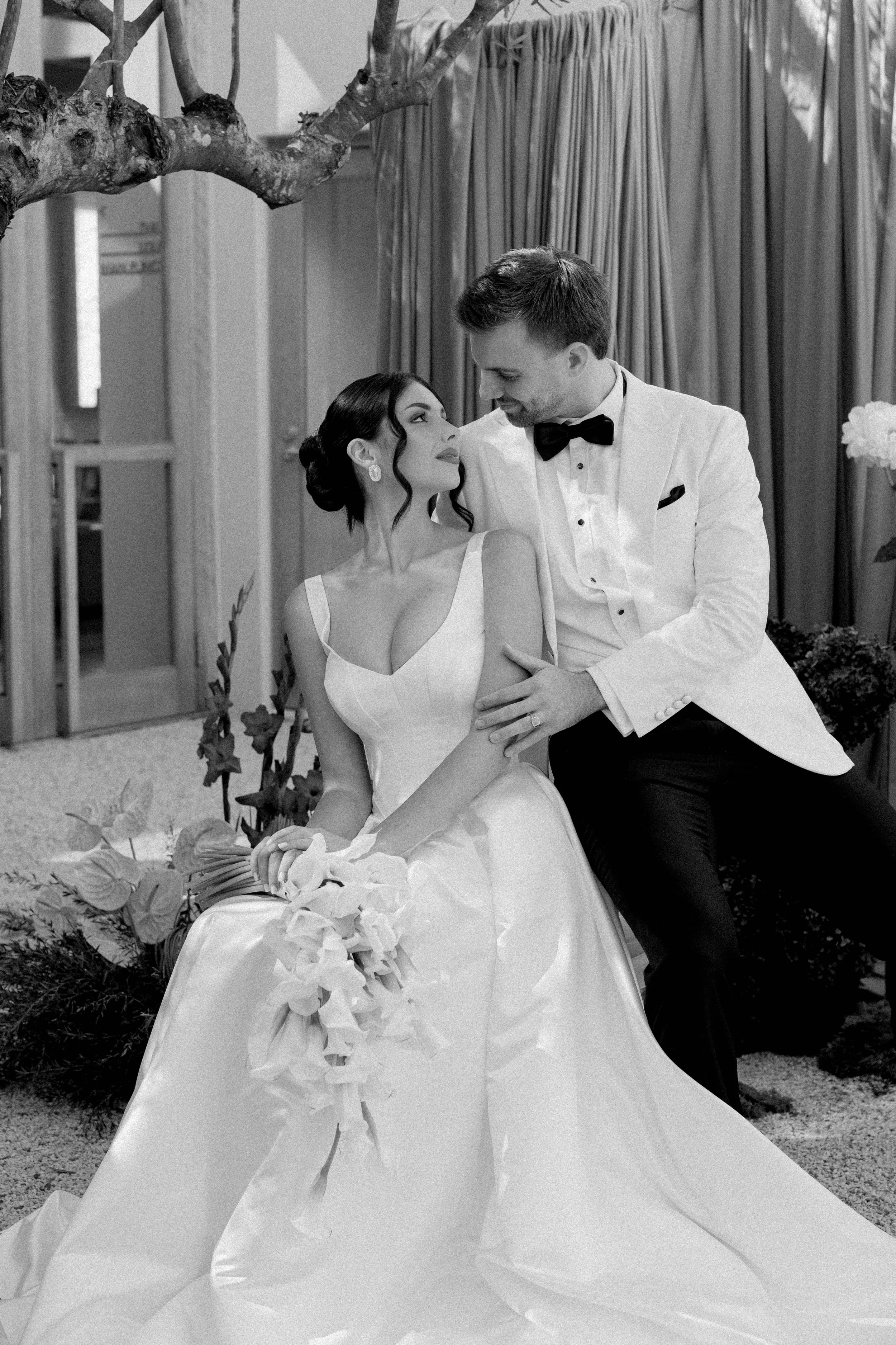 A bride and groom sharing a tender moment during their wedding, with the bride sitting and the groom leaning over her, both smiling lovingly. The setting appears formal with luxurious curtains and floral decorations.
