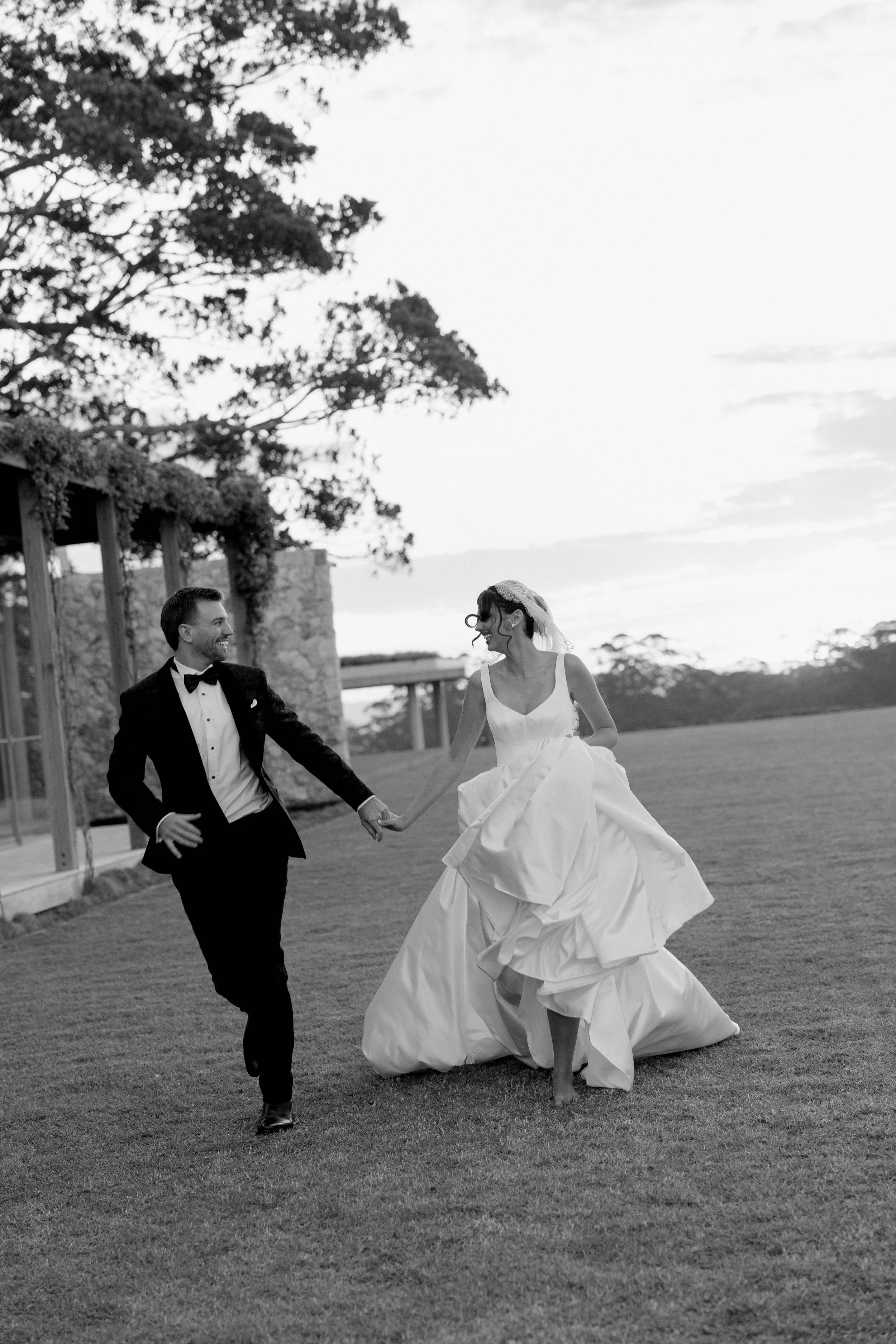 A black and white photo of a bride and groom dancing outside on a grassy area during sunset, holding hands and smiling at each other.