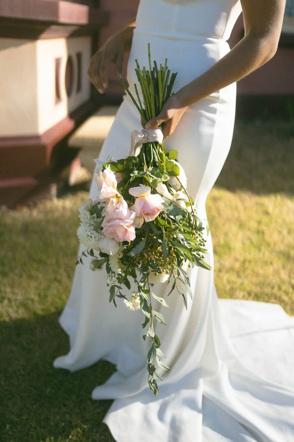 Person in a white dress holds a cascading bouquet of pink and white flowers with greenery outside on grass.