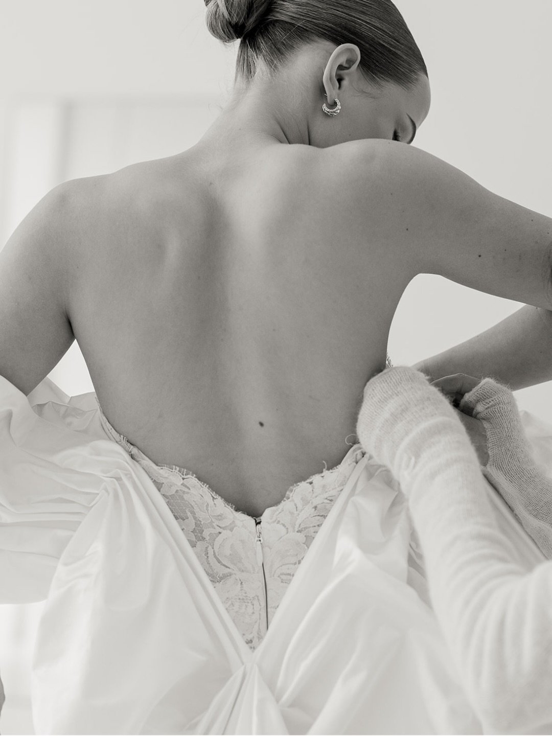 A woman with her back facing the camera, partially dressed in a white garment, receiving assistance with her dress or gown from another person who is wearing gloves.