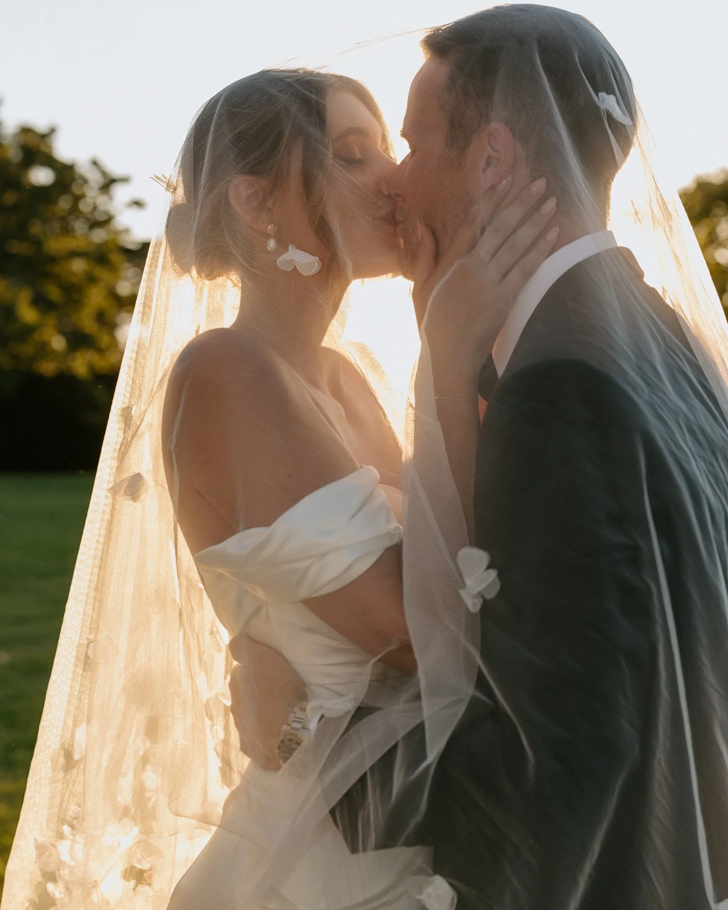 A bride and groom sharing a kiss outdoors during sunset, with the sunlight backlighting them and a veil flowing over both of them.