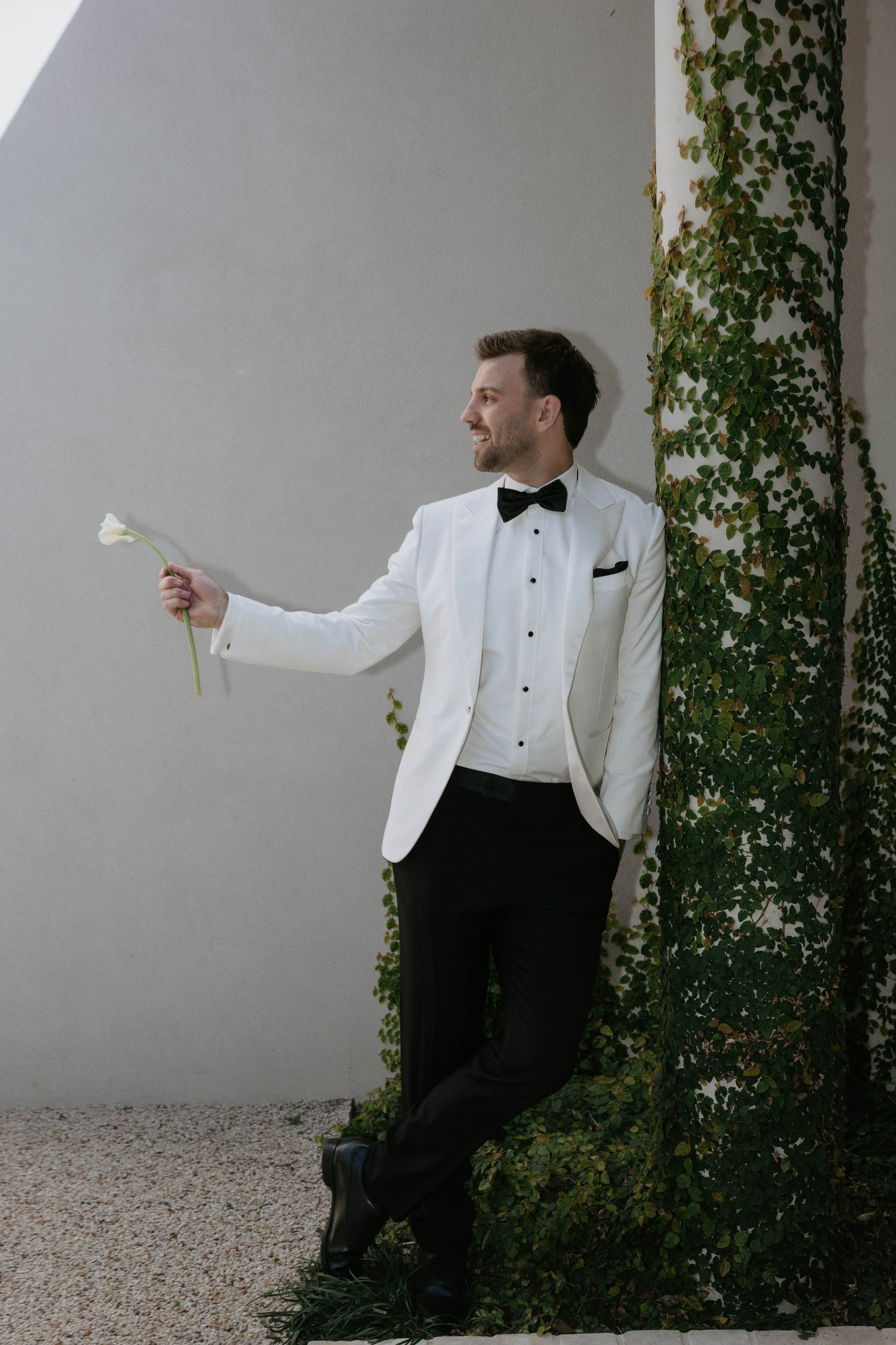 A man in a white tuxedo with a black bowtie and black pants standing against a wall covered with green ivy, holding a white flower in his right hand and smiling.