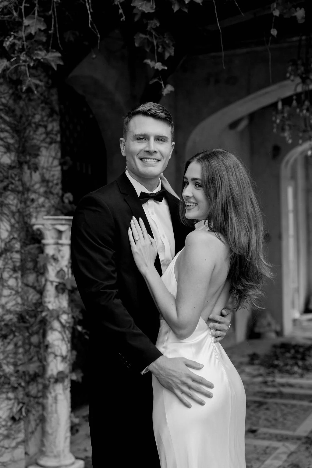 A black and white photo of a couple dressed in formal attire, smiling and embracing each other, with a decorative garden or courtyard background.
